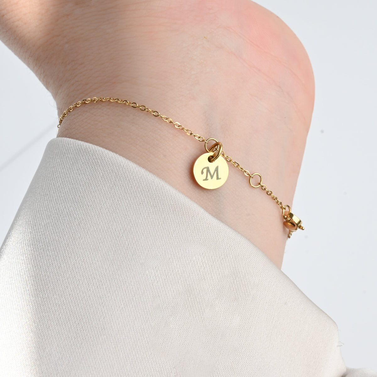 Gold bracelet with a round initial charm on a person wearing a white shirt against a light gray background