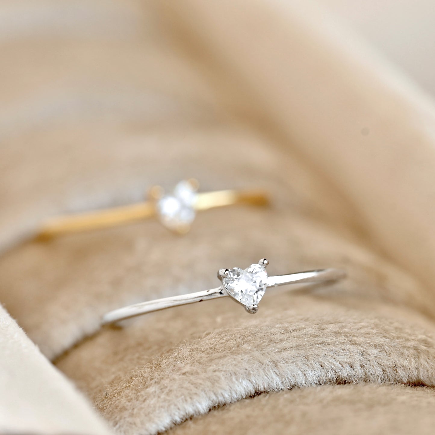 Two delicate solitaire rings with heart-shaped clear gemstones, one in silver and one in yellow gold, displayed on a beige velvet ring box.