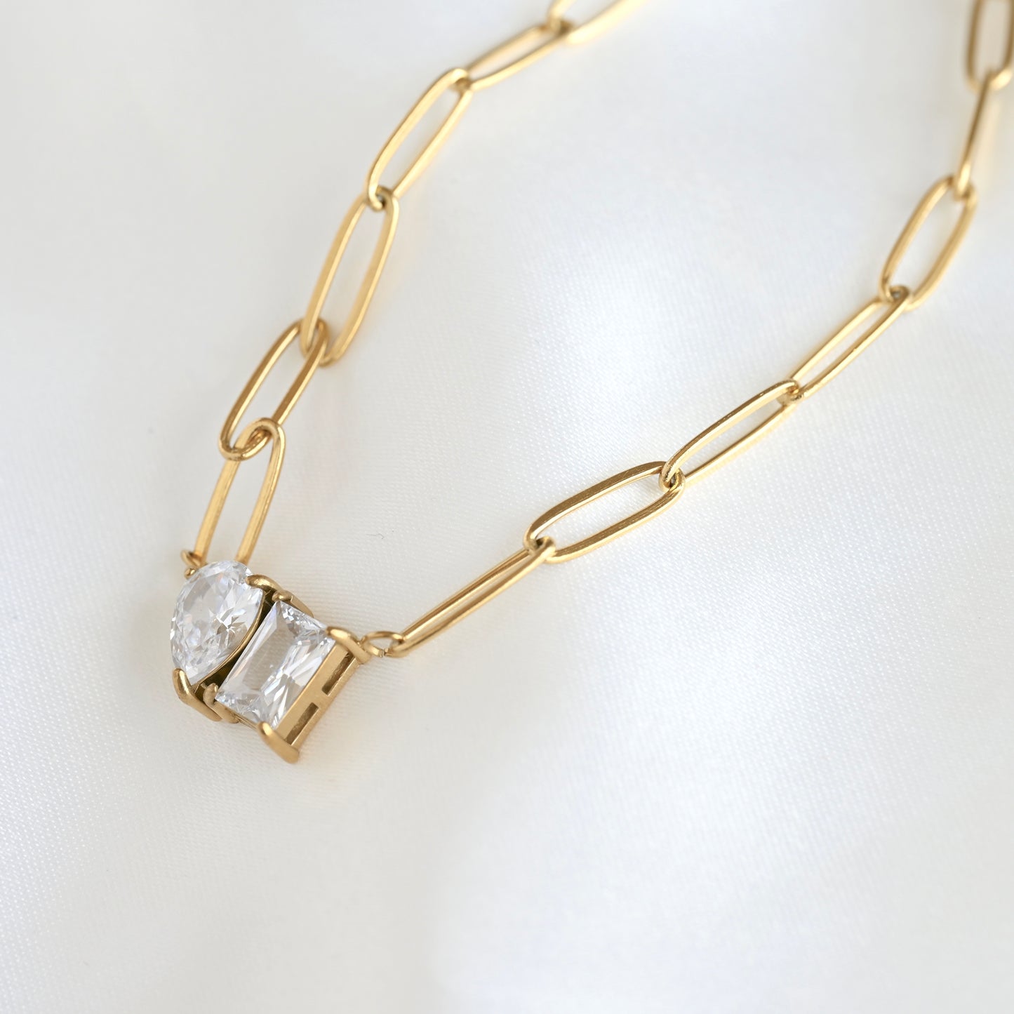 A gold paperclip chain necklace featuring a small pendant with two clear simulated diamonds in pear and rectangular shapes, displayed on a clean white fabric background.