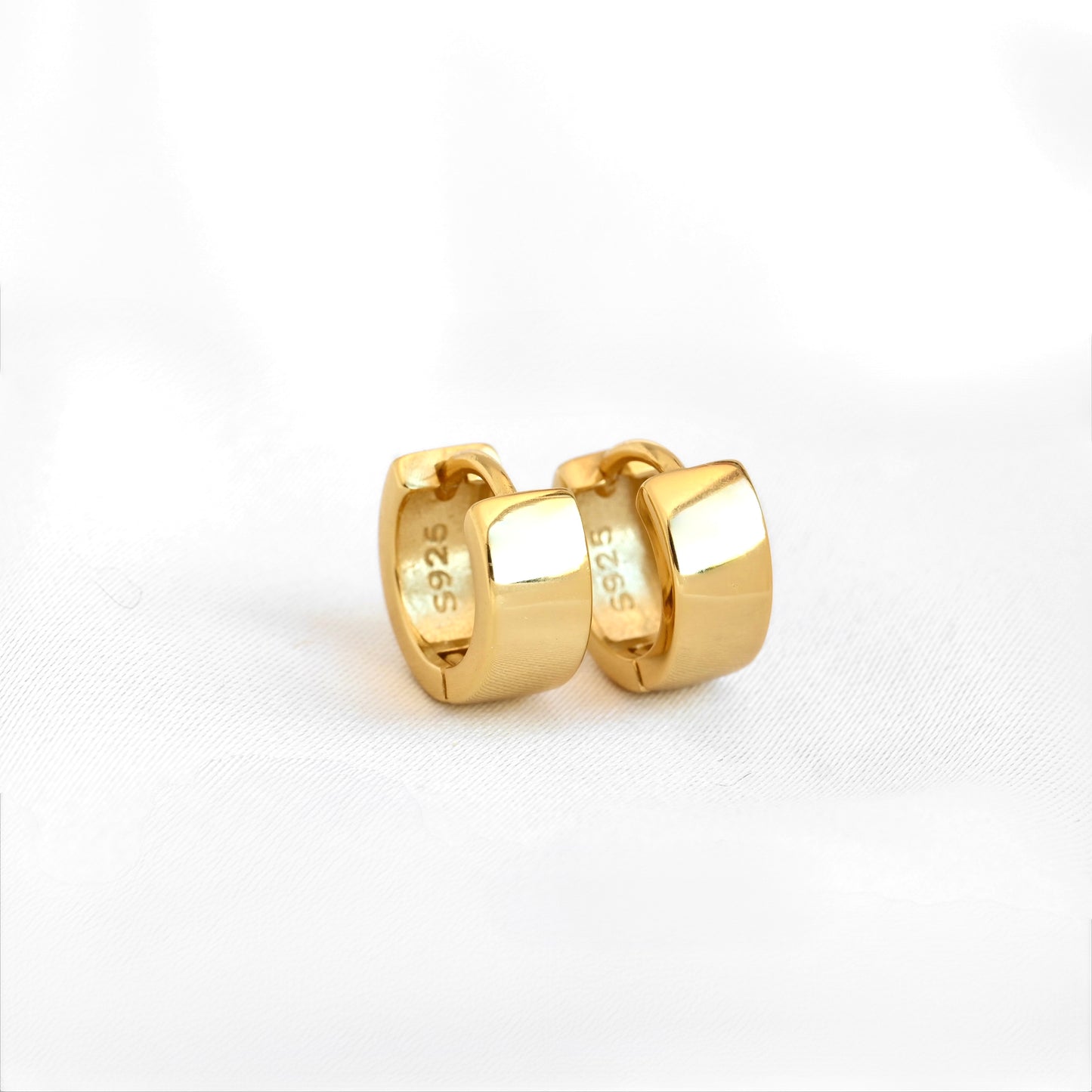 An gled front view of a pair of polished yellow gold huggie hoop earrings with a 925 sterling silver stamp on the inside, shown on a clean white textured fabric background.