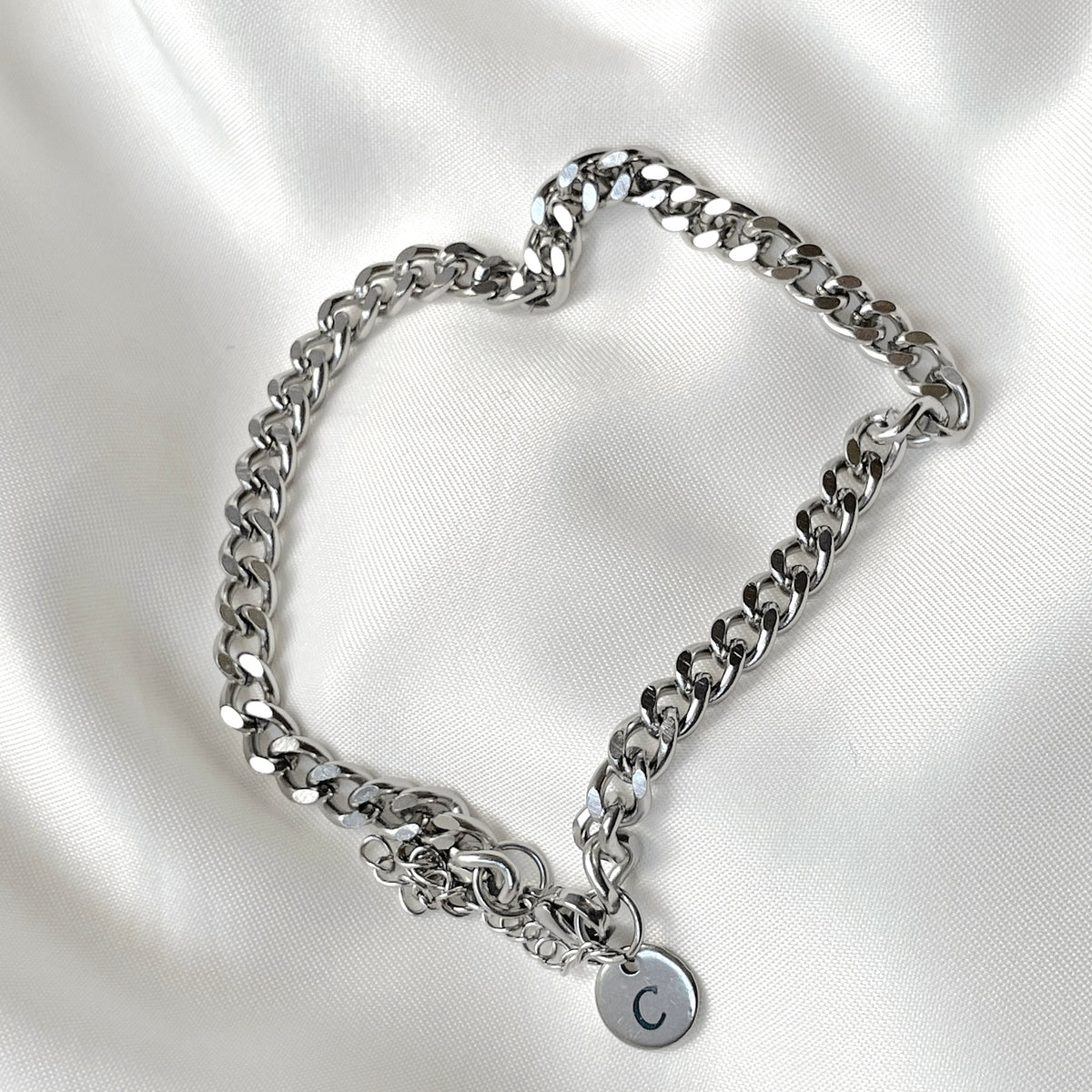 Thick silver chain ankle bracelet with a round initial charm on a white satin background