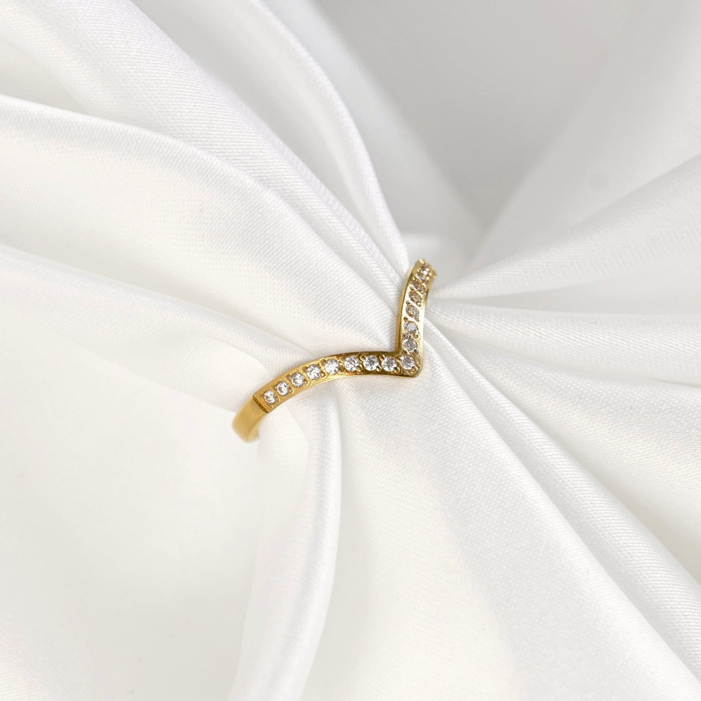 A gold wishbone ring with small clear stones, displayed on a draped white satin fabric.