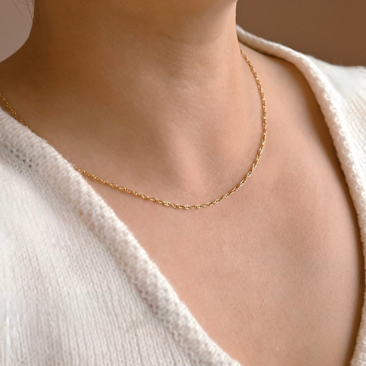 A delicate gold chain necklace worn on a person's neck, styled with a cream-colored knit top.