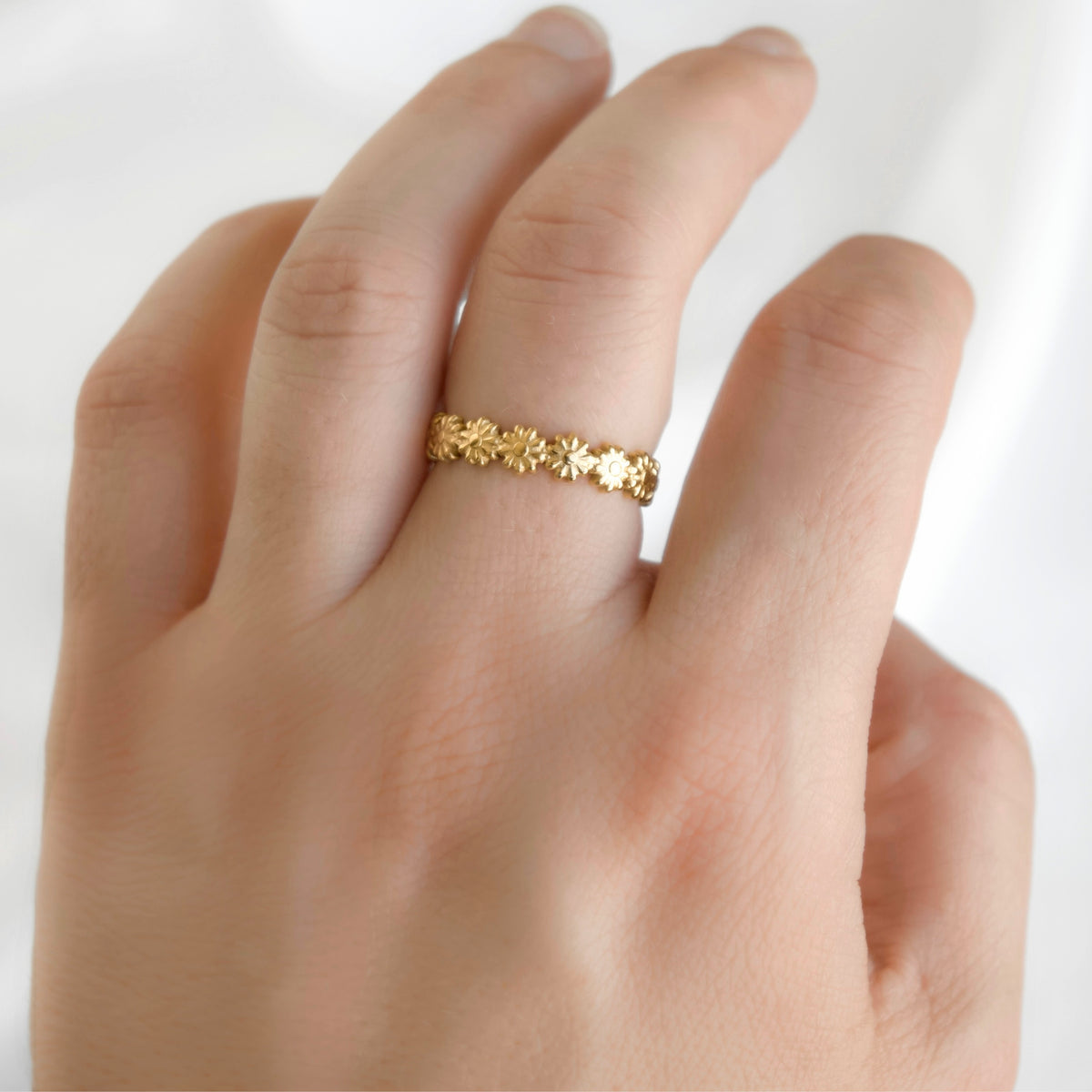 Dainty gold daisy chain band on a woman's middle finger against white background.