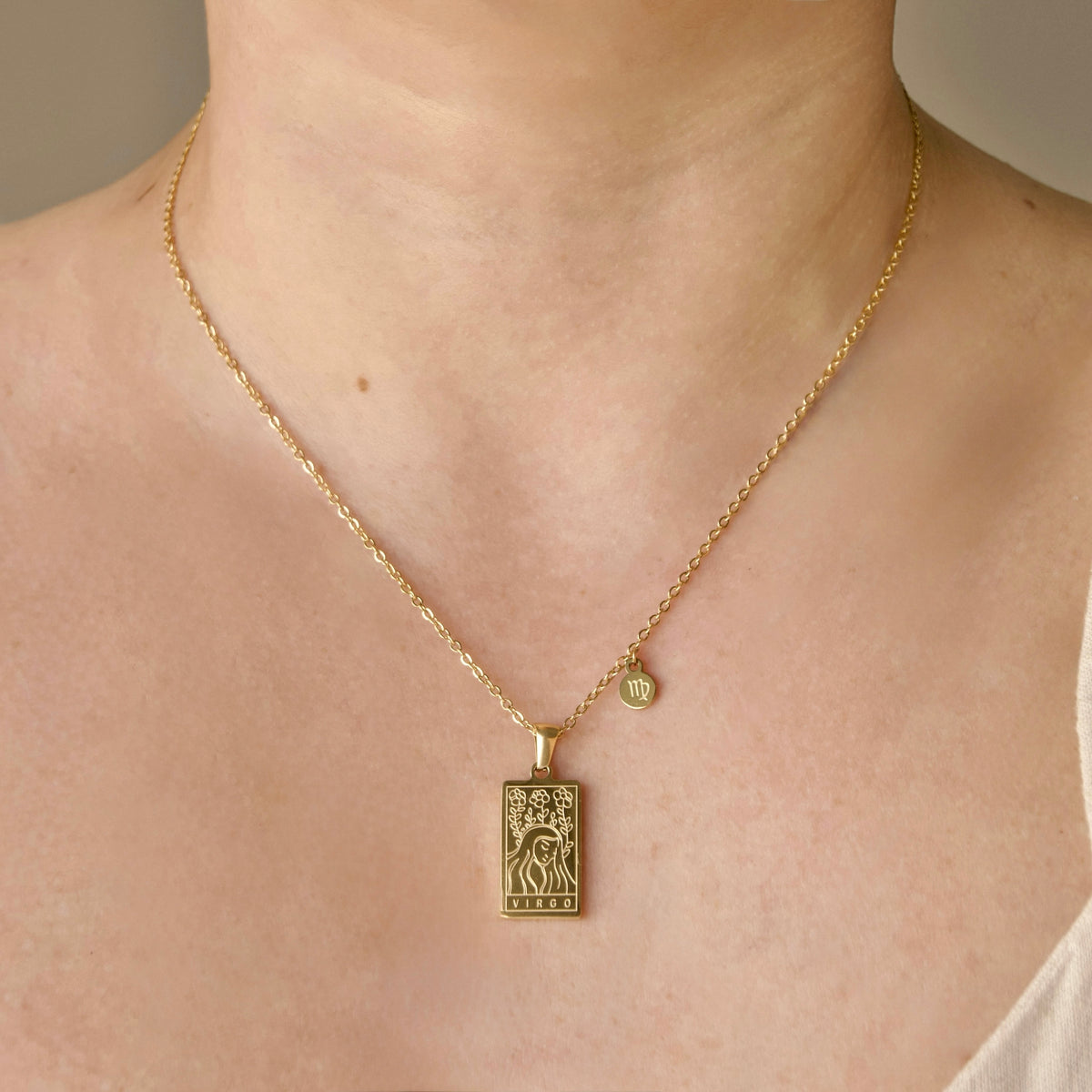 A close-up of a person's neck and chest wearing a delicate gold chain necklace with a rectangular engraved zodiac pendant and a small round charm.