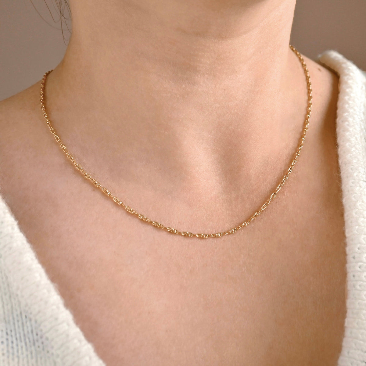 A close-up of a person's neck and upper chest wearing a delicate gold chain necklace with a small mariner chain link design, paired with a white knit top.