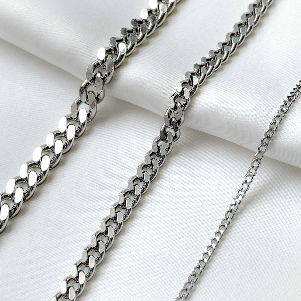 Three silver cuban bracelets in various thicknesses on a white fabric background