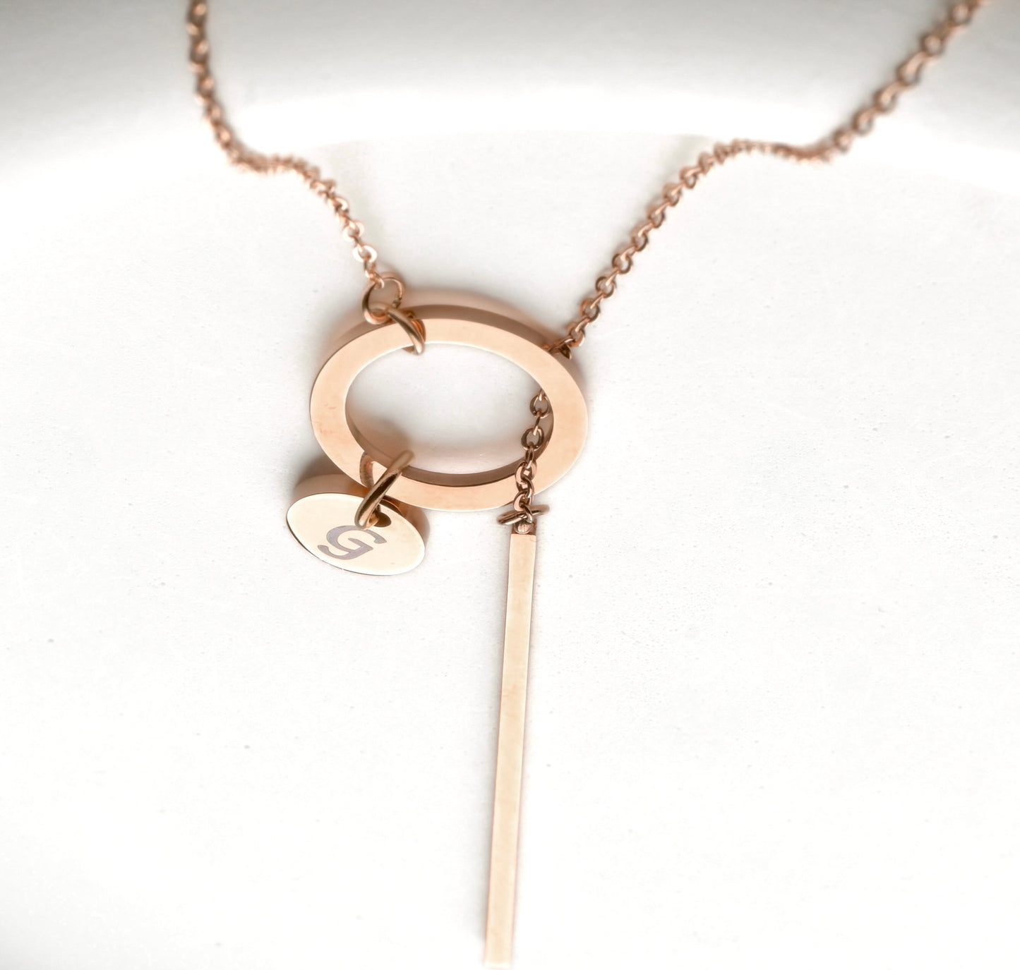 Top view of rose gold necklace with a circle, 3d bar and a small disc initial charm on a white background