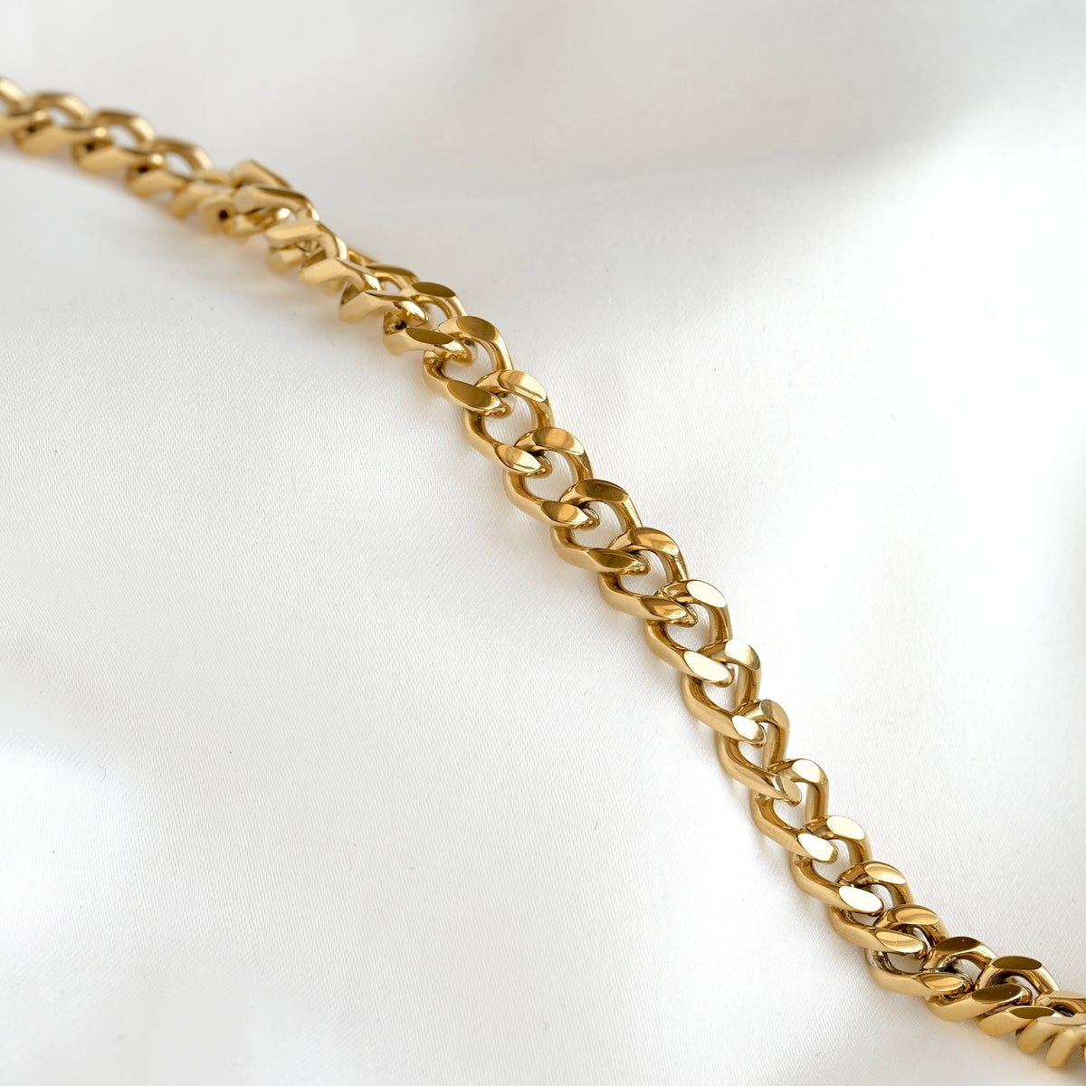 A close-up of a polished gold chain with interlocking curb-style links, laid diagonally across a soft white fabric background.
