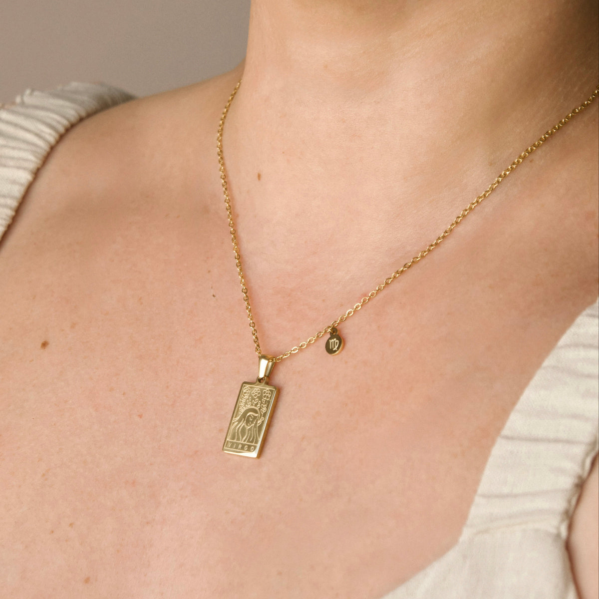 A gold pendant necklace worn by a model. The necklace features a rectangular engraved pendant with virgo depiction and text under and a small round charm with virgo symbol engraving on a delicate gold chain.