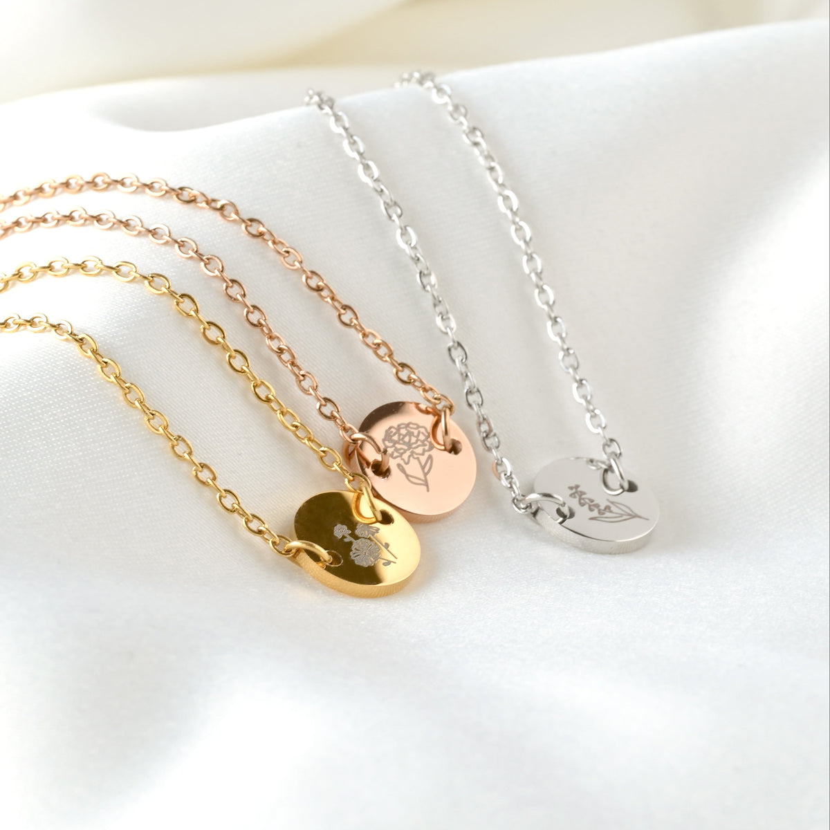 Three bracelets with round birth flower charms in gold, rose gold and silver on a white surface