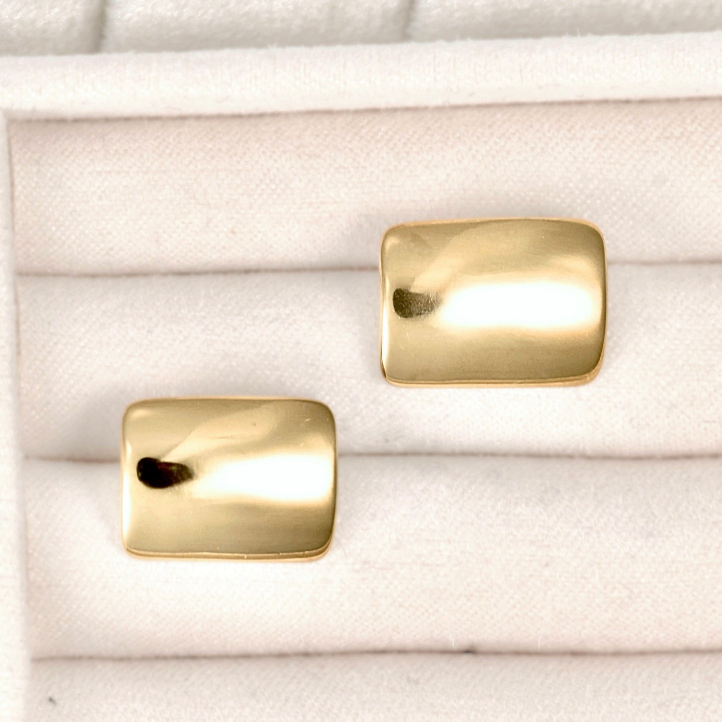 A pair of gold-toned rectangular stud earrings with a polished, slightly rounded finish, displayed on a white textured ring box.