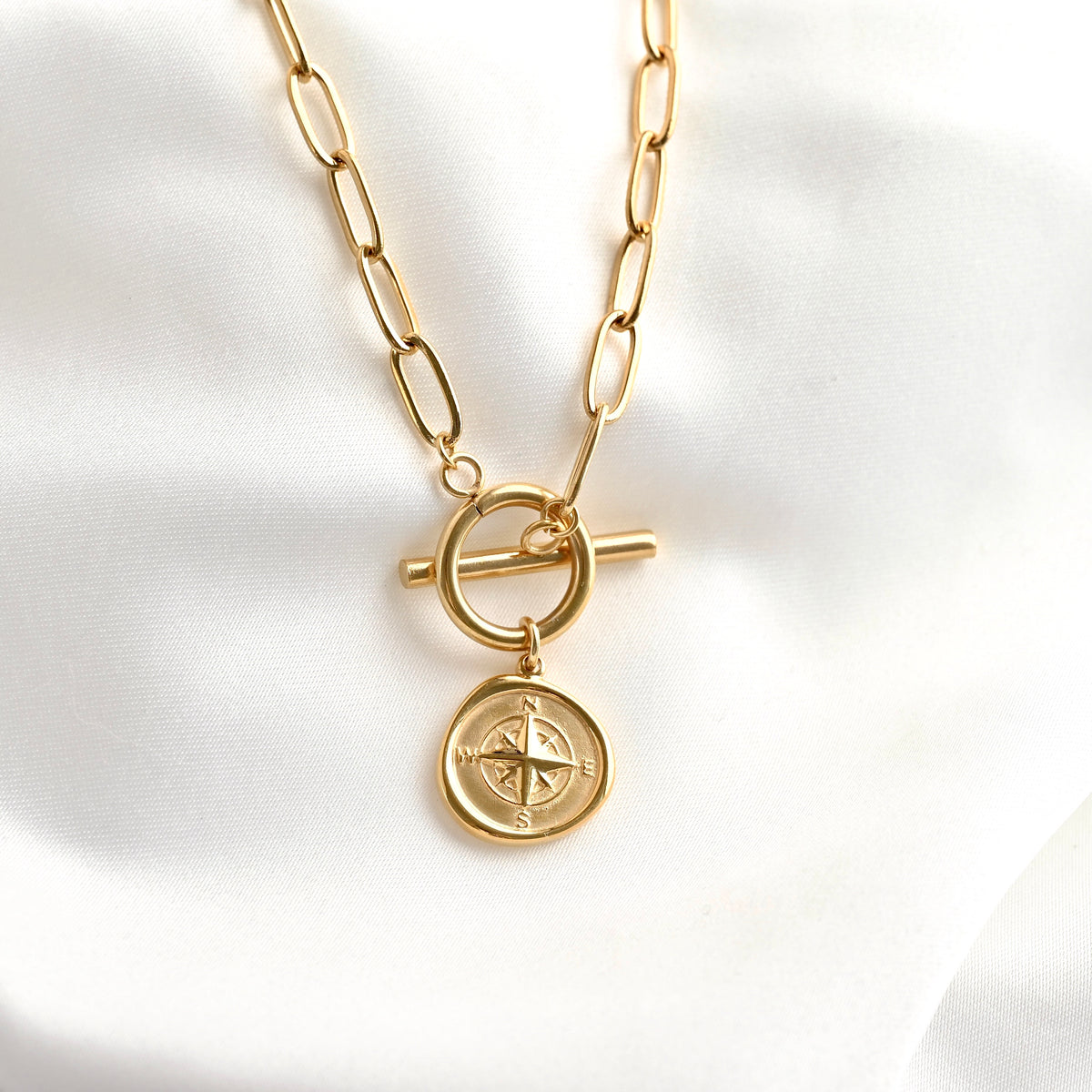 A gold-toned chain necklace with a toggle clasp and a round compass pendant, displayed on a soft white fabric background.