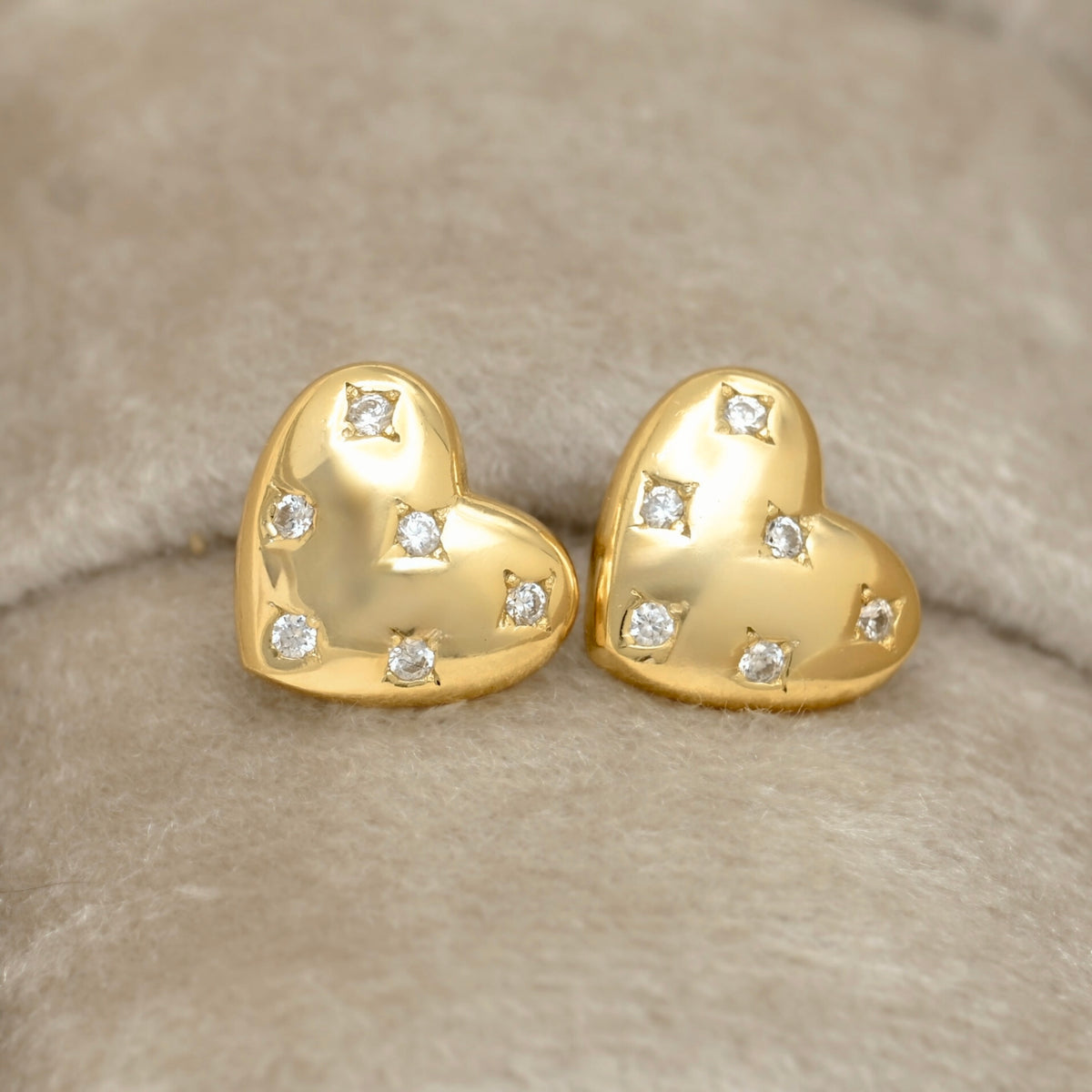 A pair of gold heart-shaped stud earrings, each adorned with small clear round CZ stones in a star setting, displayed on a soft beige textured surface.