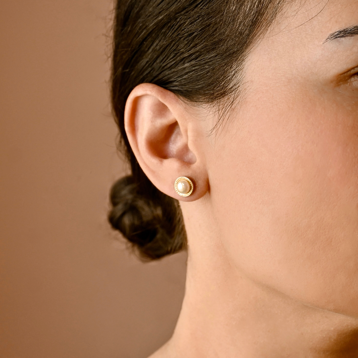 A close-up of a woman's ear wearing a small round gold stud earring with a white pearl center, against a warm brown background.