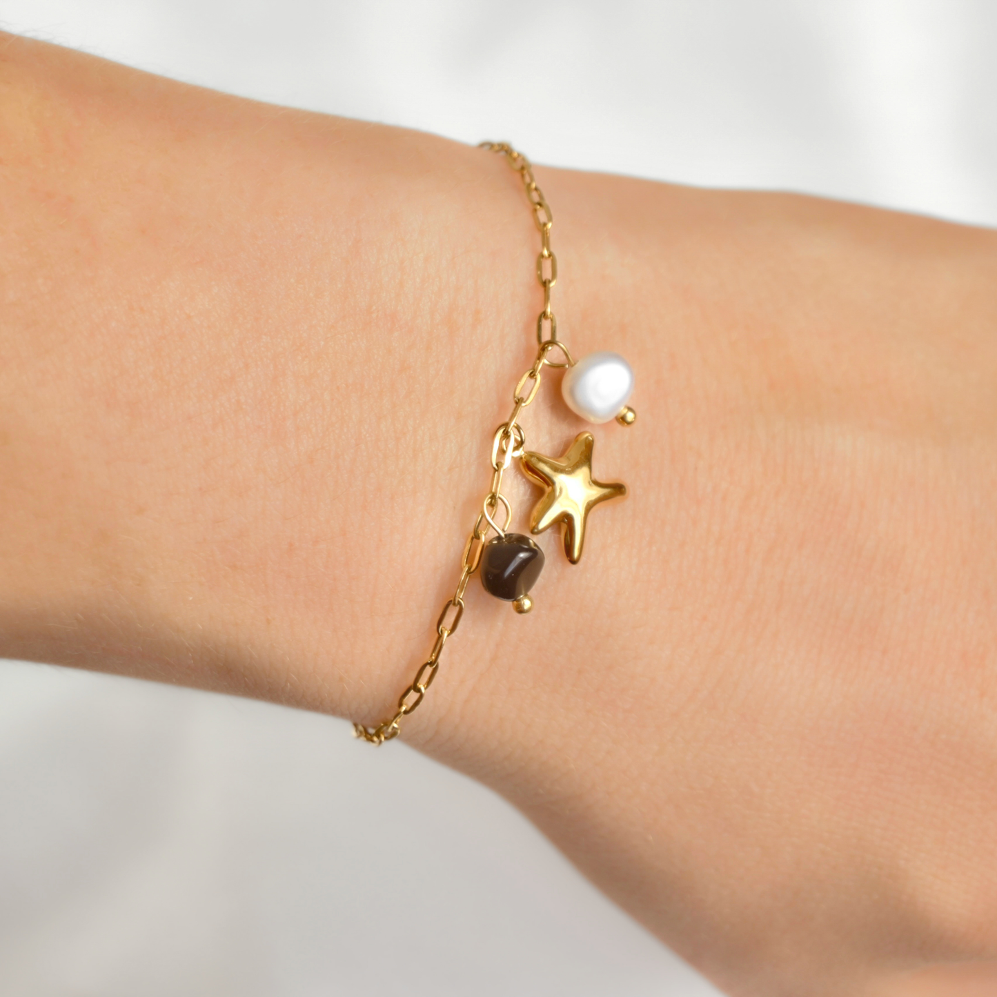 A delicate gold chain bracelet with a sea star charm, a white pearl, and a small black stone charm, shown worn on a person's wrist.