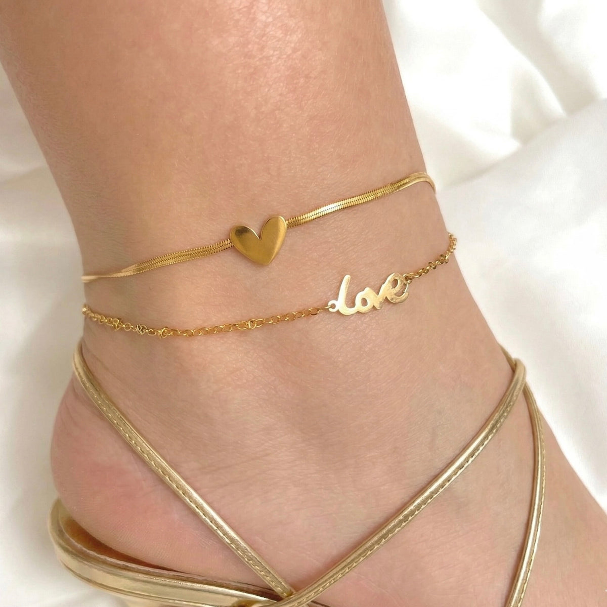 Layered gold anklet with heart and 'love' charm on a person's ankle wearing elegant gold strappy sandals.