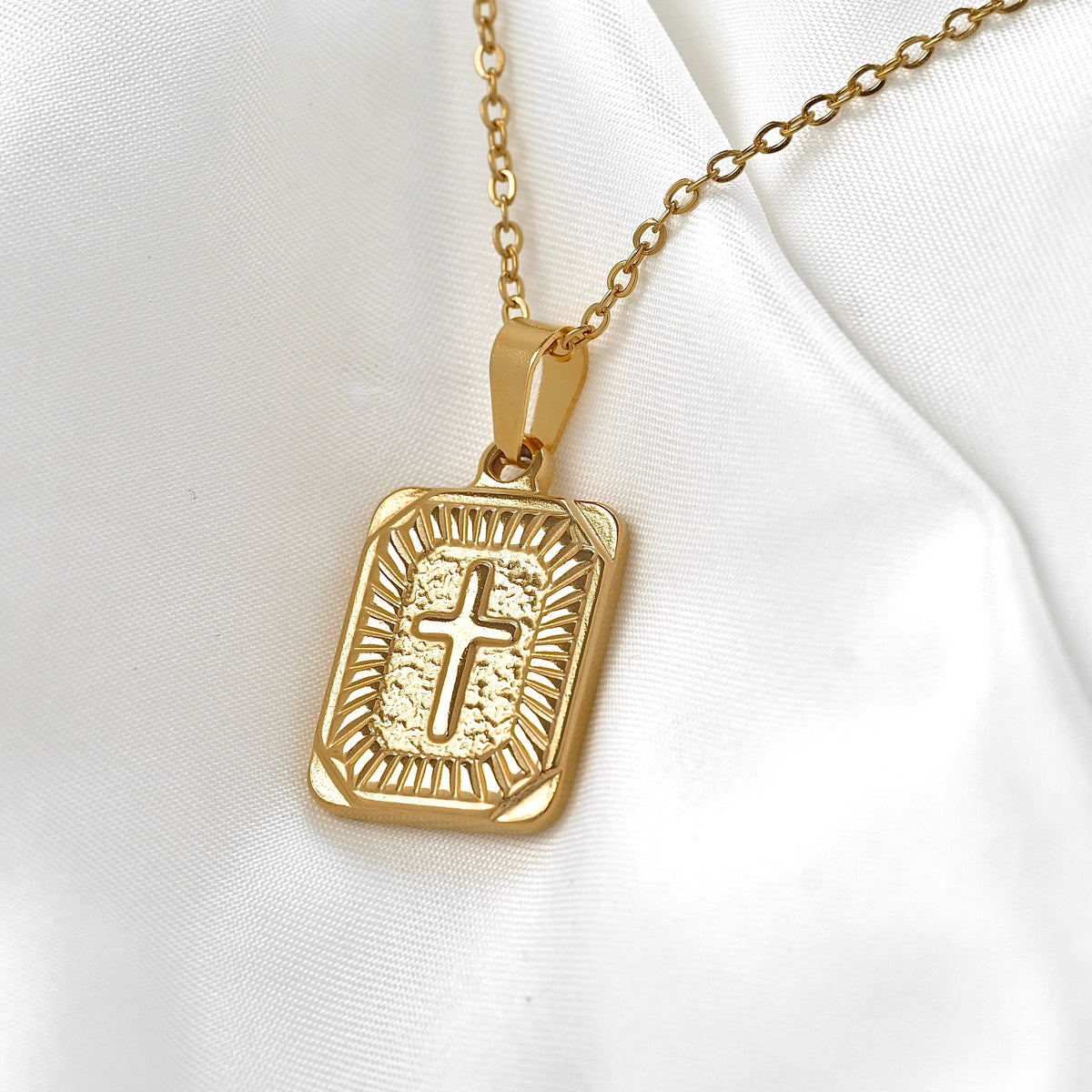 A gold rectangular pendant necklace featuring an engraved cross in the center, displayed on a soft white satin fabric background.