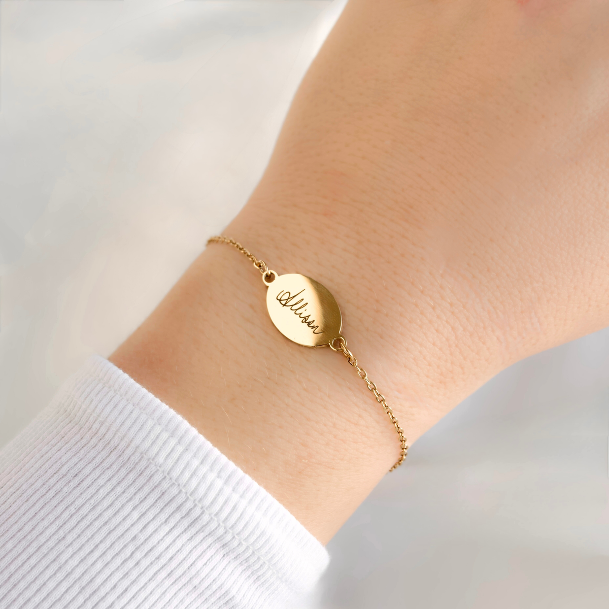 A delicate gold chain bracelet with a round engraved disc charm reading 'Alicia', worn on a person's wrist over a white ribbed cuff.