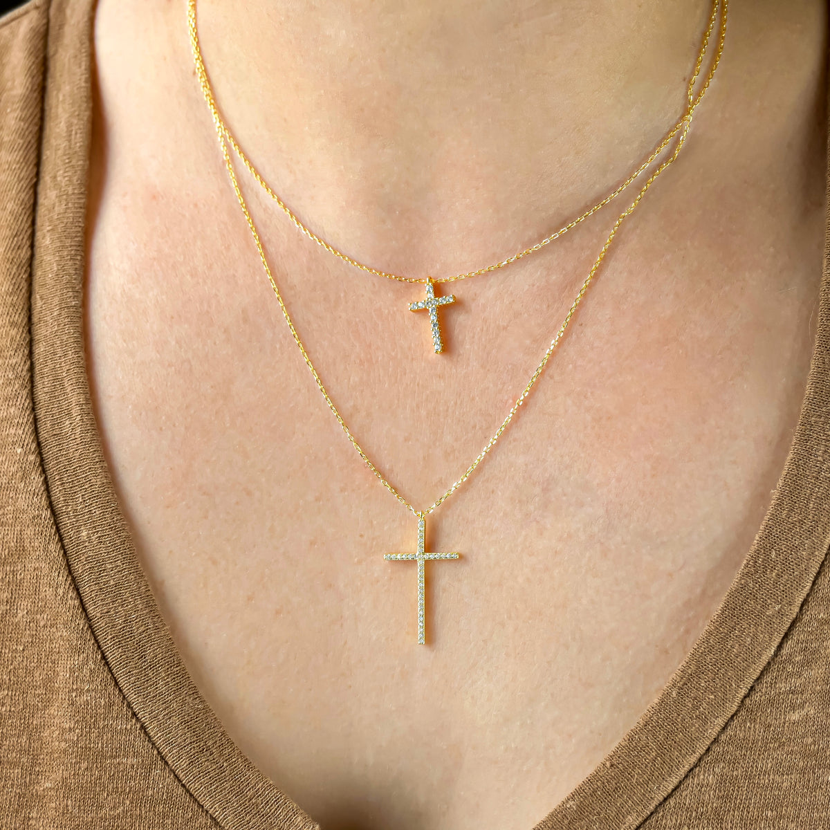 Two gold cross necklaces, one with medium size and one with large cross charm on a person wearing a brown top.