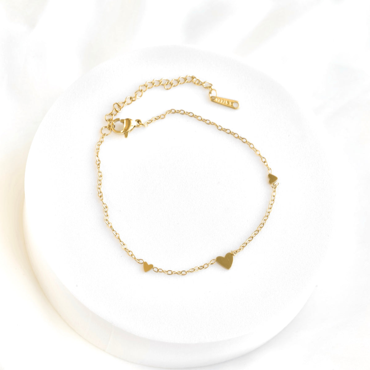 A delicate gold chain bracelet with three small flat heart charms, displayed on a round white pedestal against a soft white background.