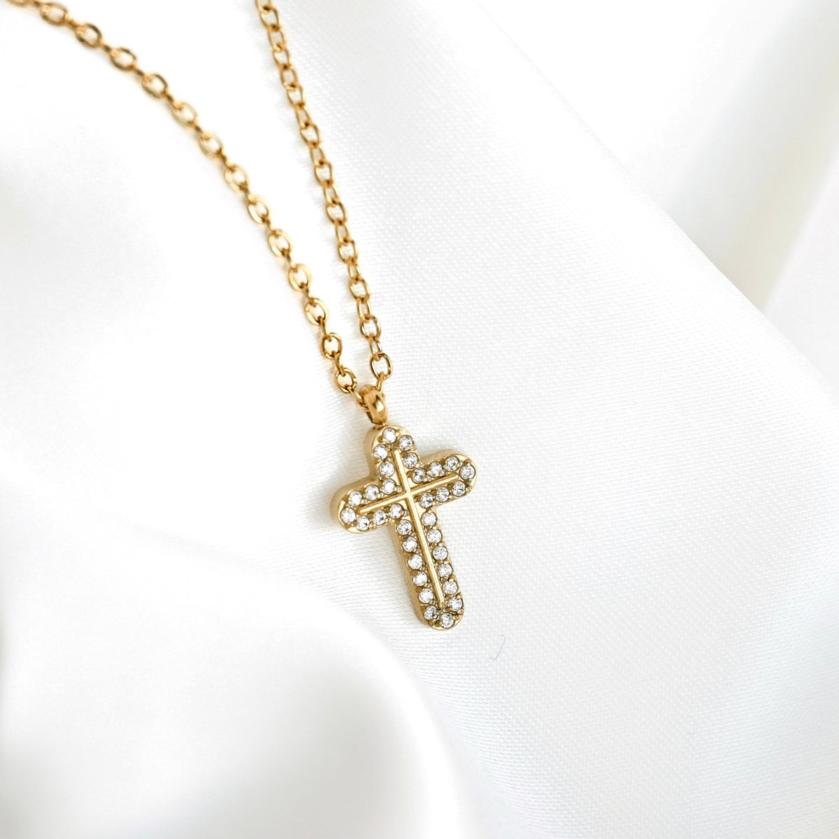 A gold cross pendant necklace with small clear rhinestones, resting on a draped white fabric background.