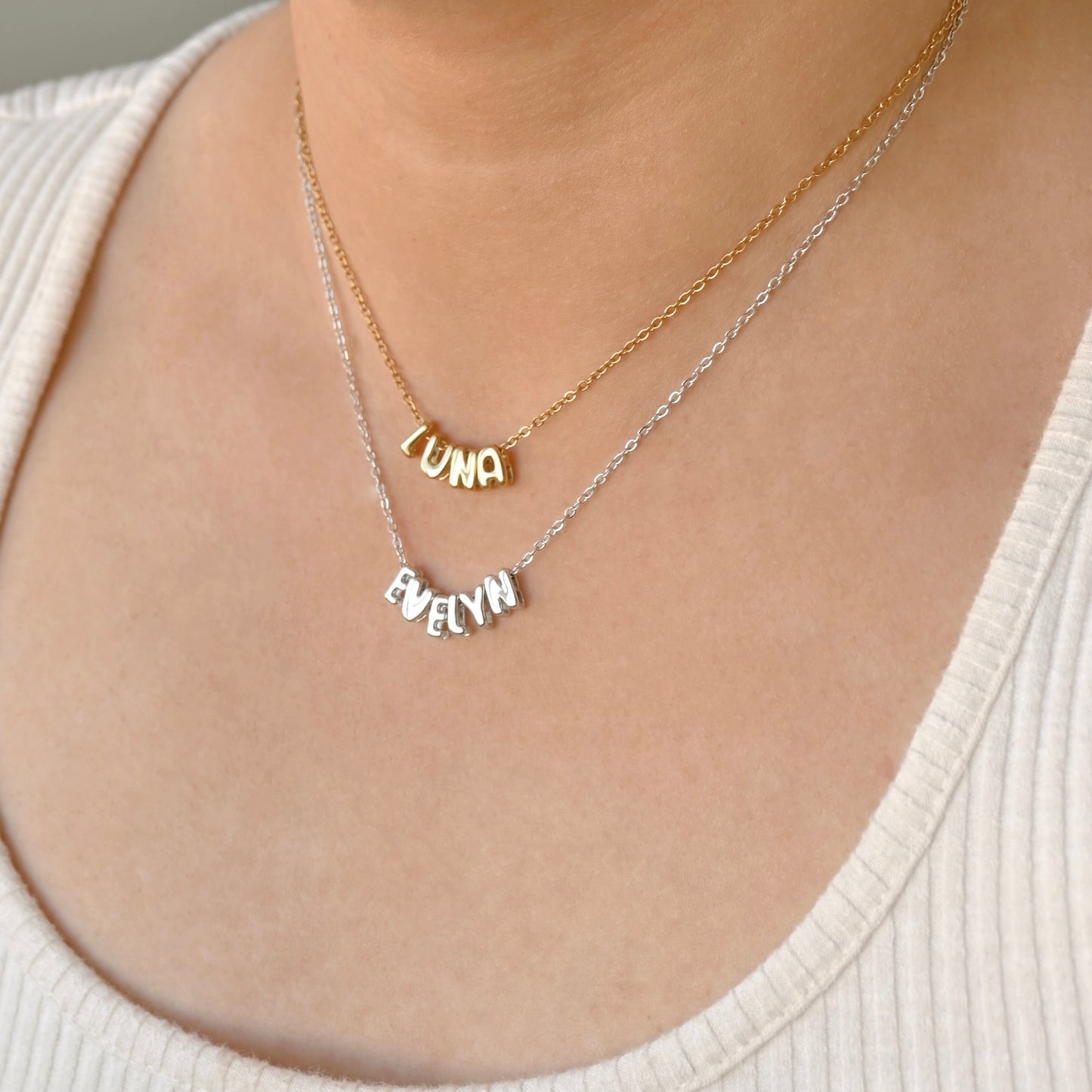 A close-up of a person wearing two delicate necklaces: a gold chain with a 'LUNA' nameplate and a silver chain with a 'EVELYN' nameplate, both displayed on the chest over a cream ribbed top.