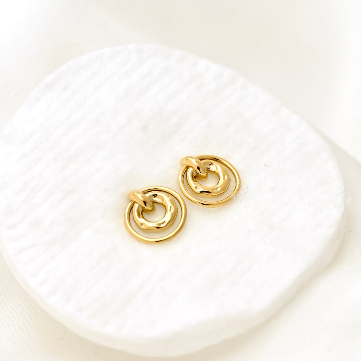 A pair of gold-toned double-ring hoop earrings resting on a textured white display pad against a soft white background.