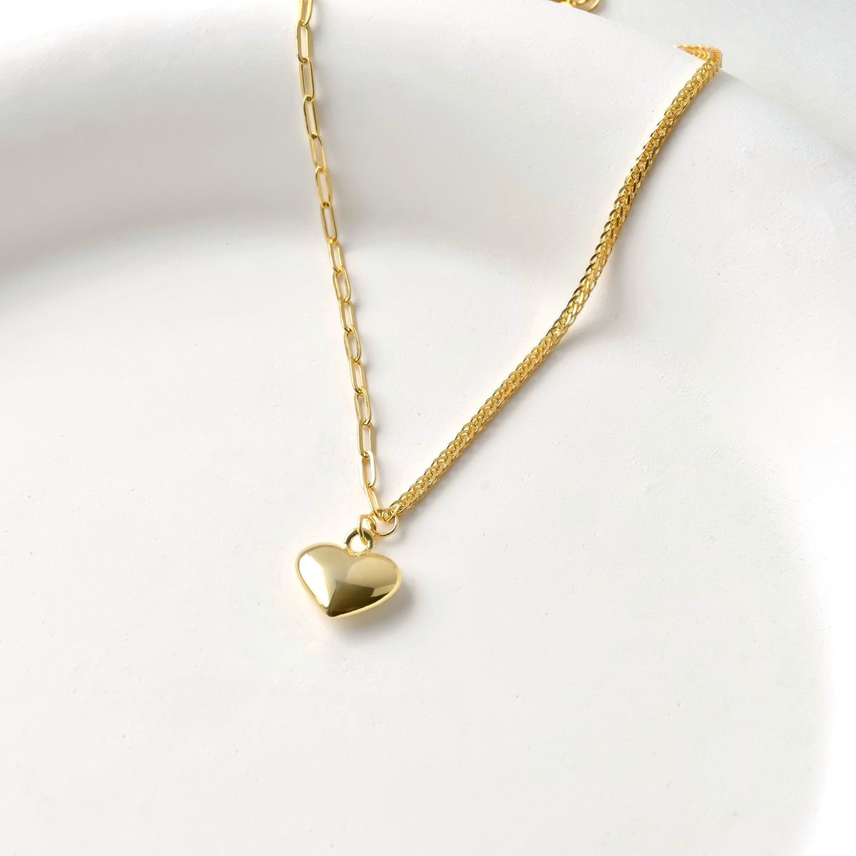 GOld puffed heart charm hanging between a dainty paperclip chain and a wheat chain on a white background