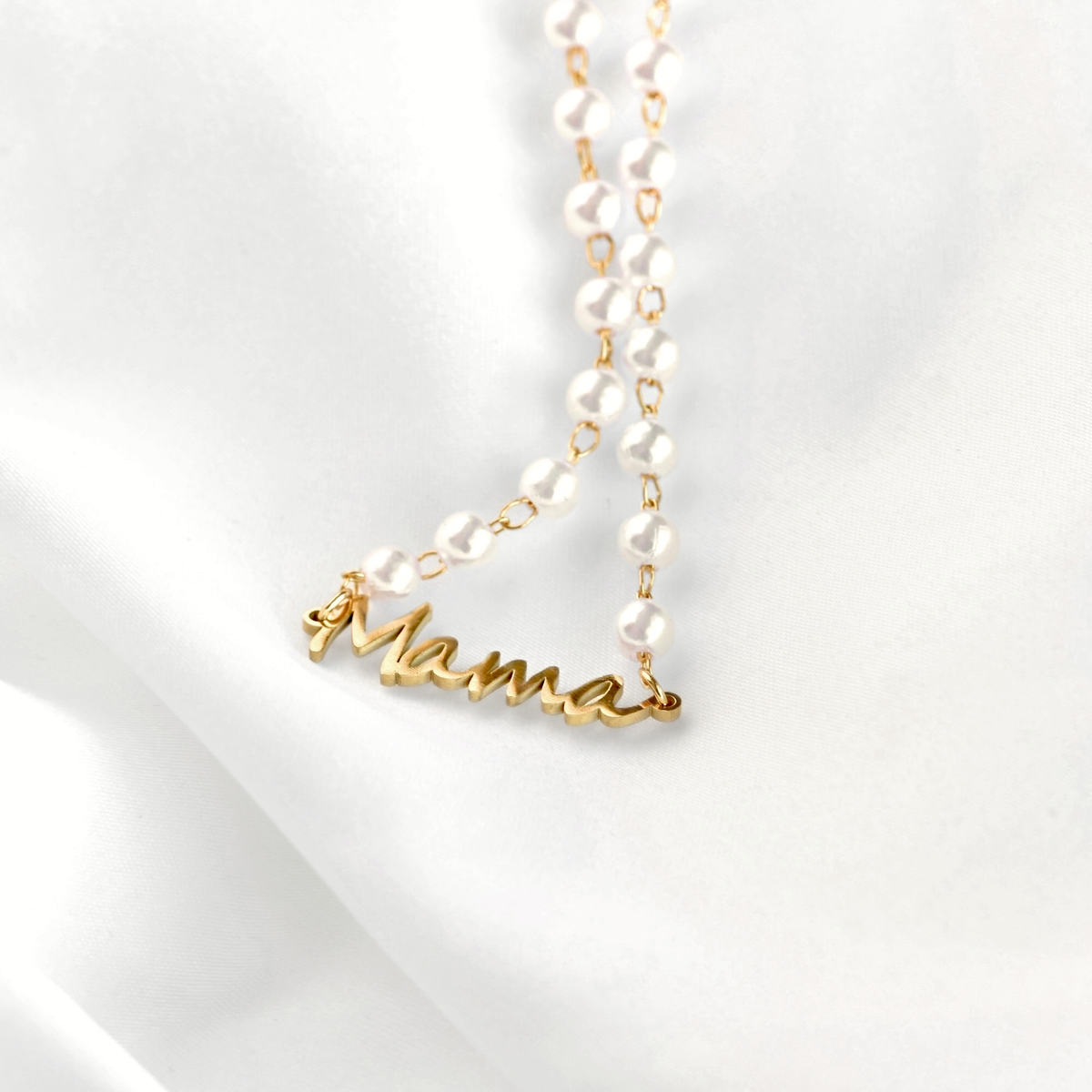 A delicate necklace featuring white faux-pearl beads on a gold chain, with a gold script pendant spelling 'Mama', displayed on a soft white fabric background.