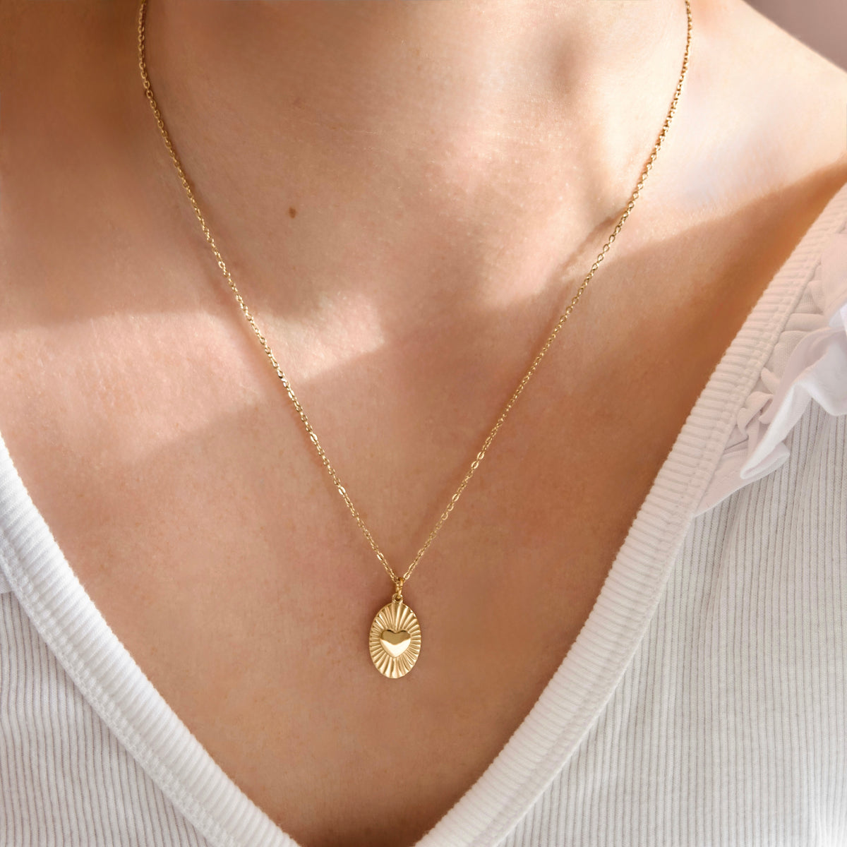 A delicate gold chain necklace with a small oval pendant featuring a sunburst design and a tiny heart in the center, worn by a model in a white ribbed top.