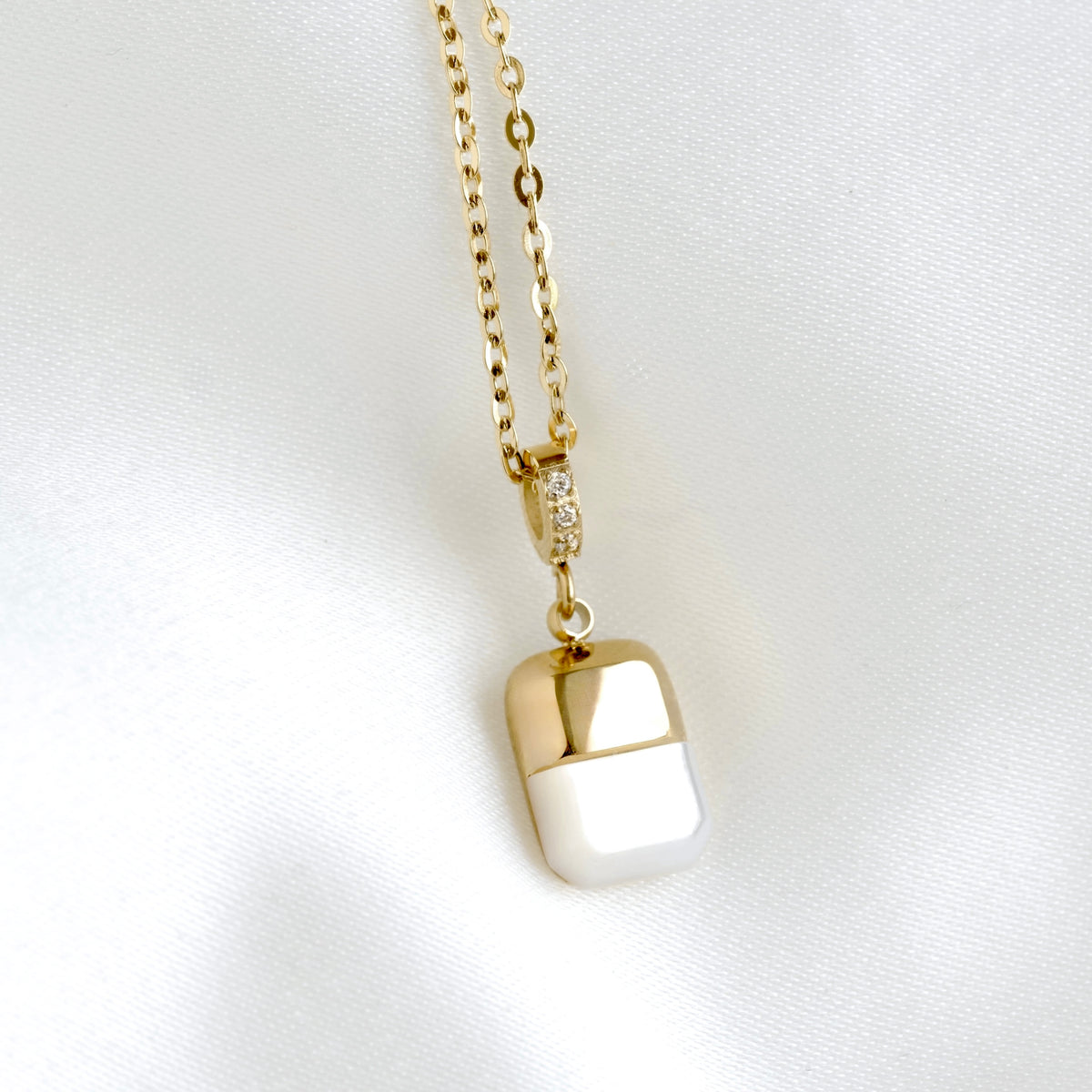A gold chain necklace with a capsule-shaped pendant, featuring a polished gold top and bottom half separated by a white center, displayed on a white satin fabric background.