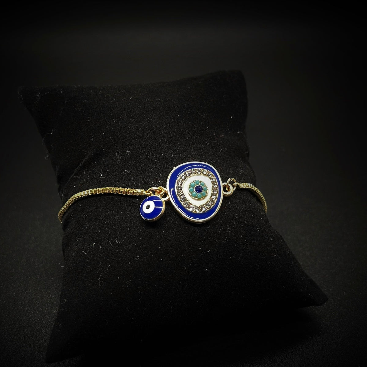Bracelet with a large evil eye charm, a tiny enamel evil eye bead and a thin box chain, on a black cushion against a dark background