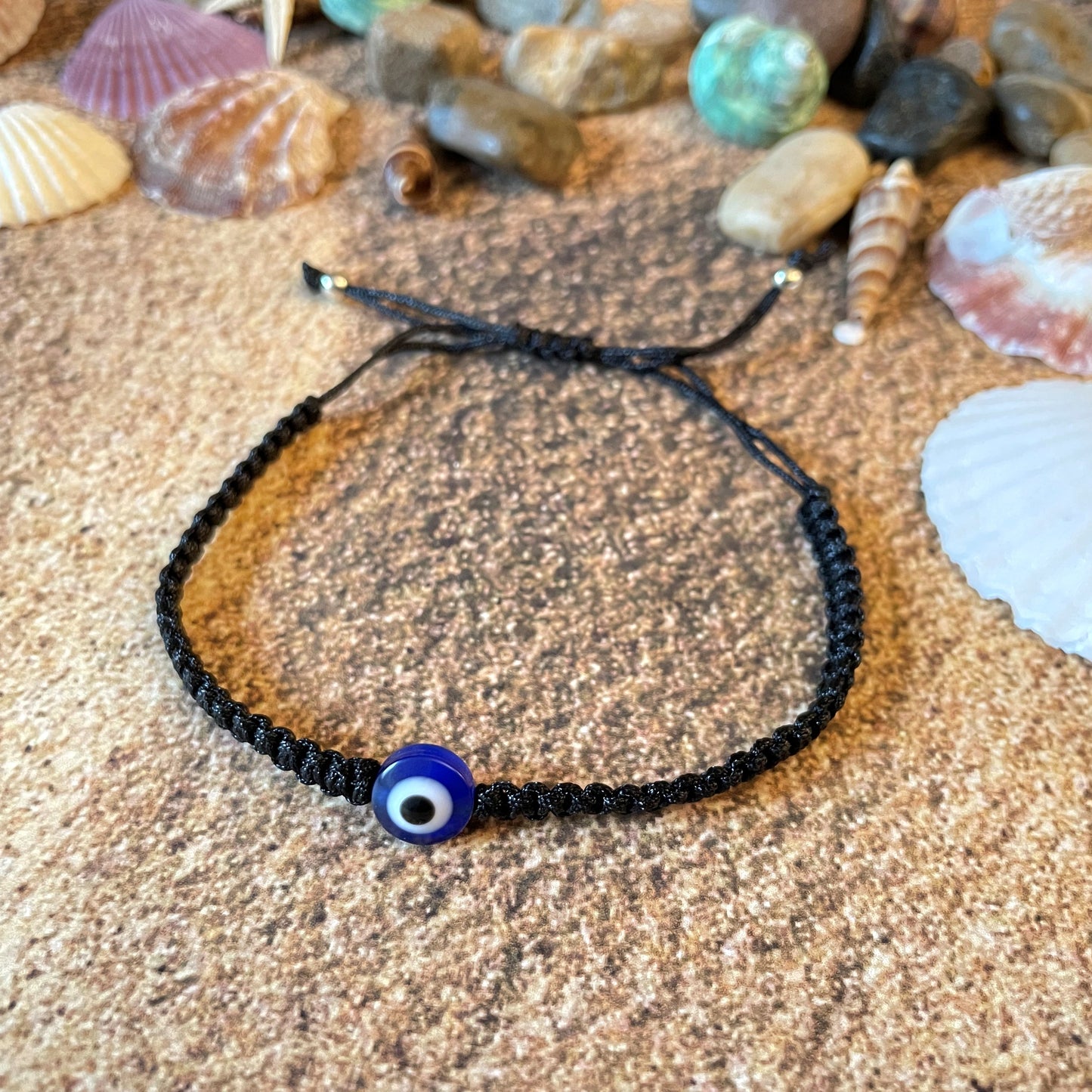 Black macrame bracelet with a blue evil eye charm on sand with seashells