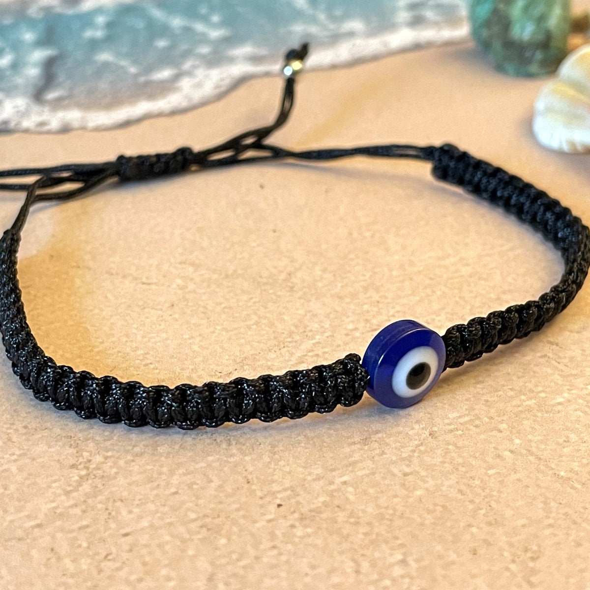Black braided bracelet with a blue evil eye bead on a sandy surface near water.