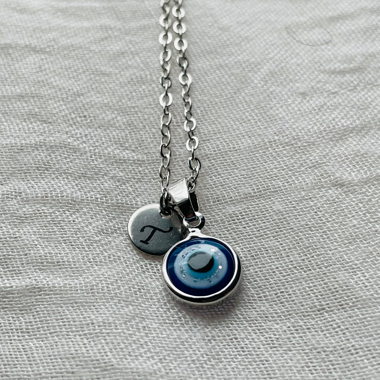 Round evil eye necklace with a small round initial disc on a textured fabric background