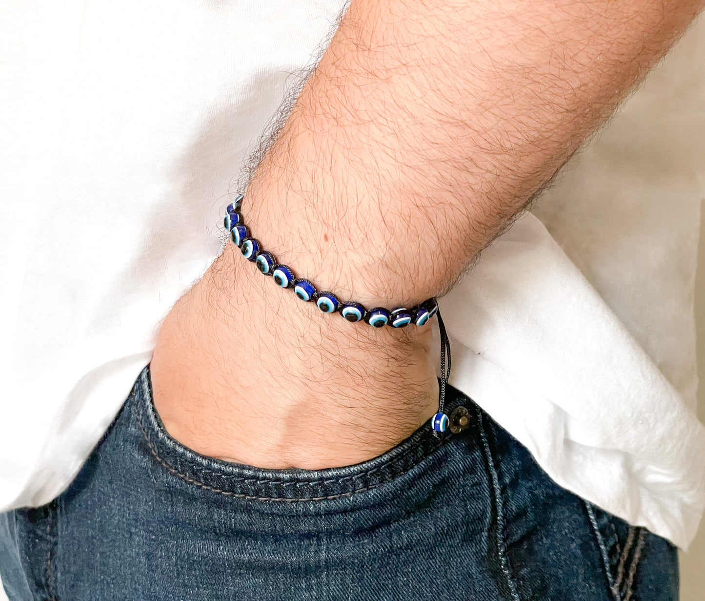 Close-up of a wrist wearing a blue evil eye beaded bracelet braided with black thread, with a white shirt and jeans in the background.