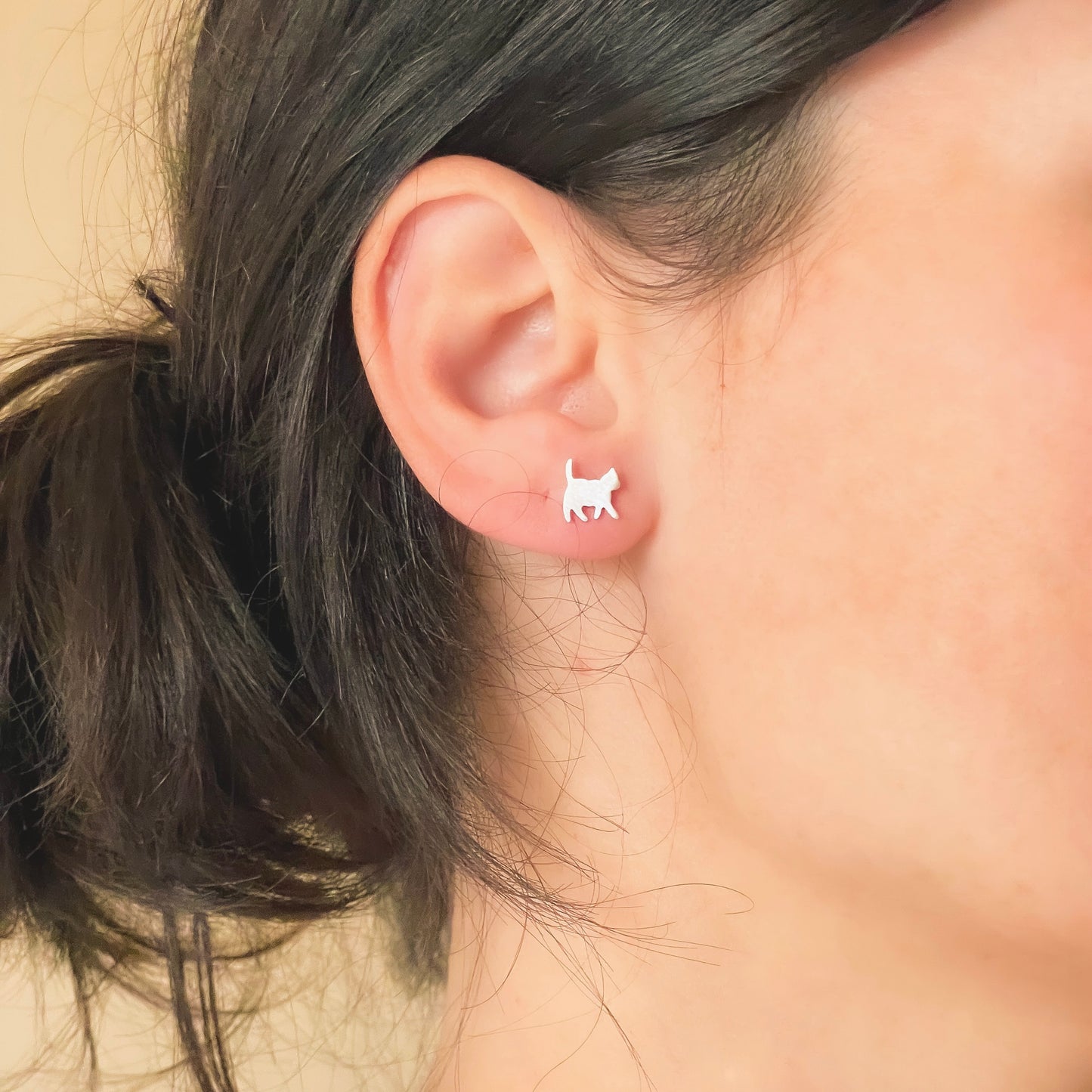 Close-up of an ear wearing small silver cat earrings with a neutral background.