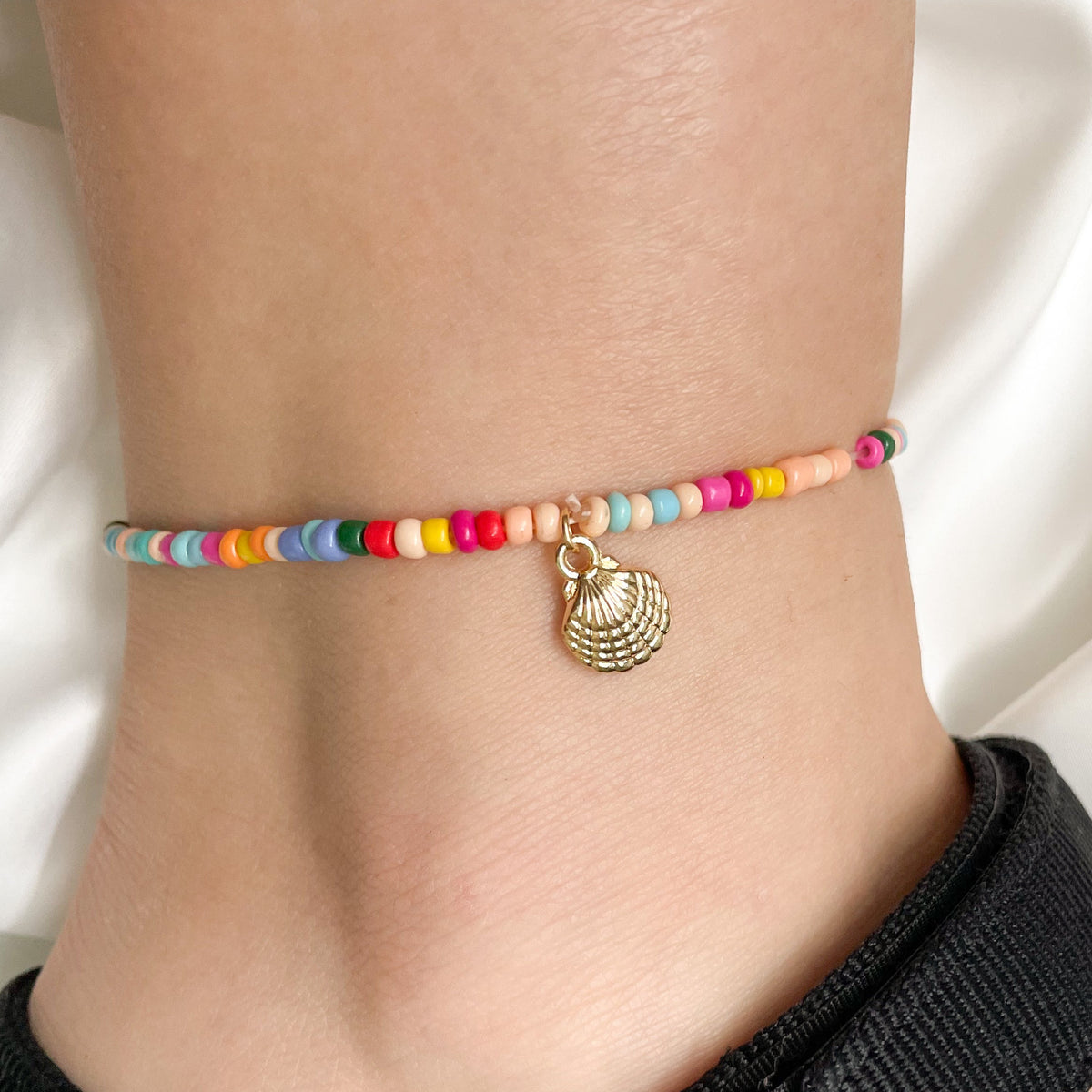 Colorful beaded anklet with a gold seashell charm on a person's ankle against a white background