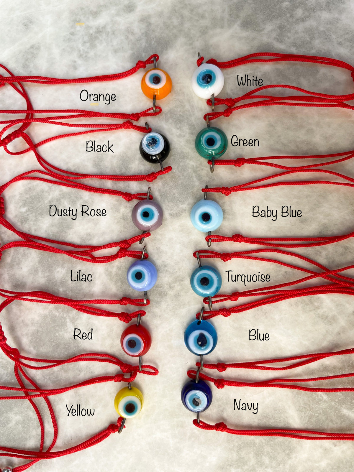 Set of 12 red string bracelets with colorful evil eyes on a gray marbled surface.