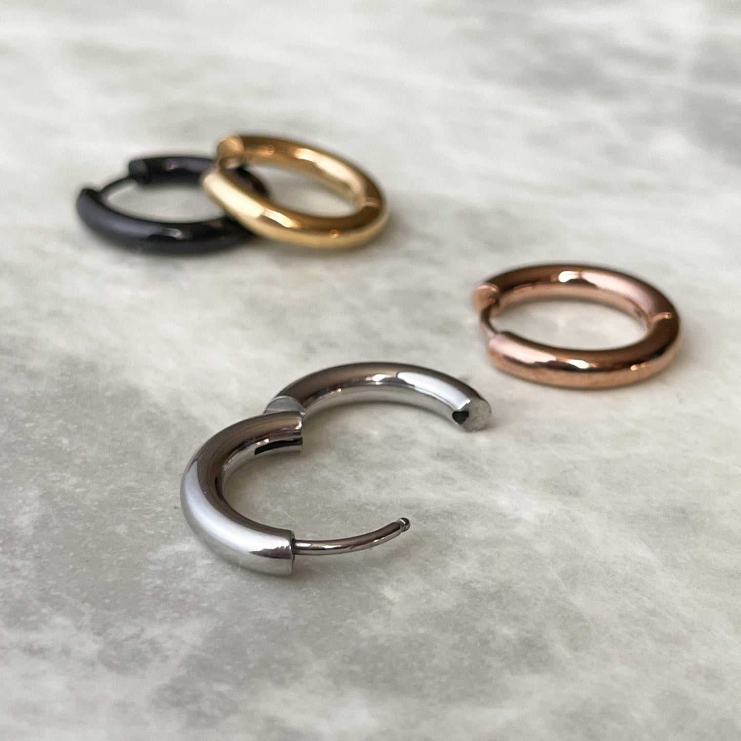 Close-up view of four small hoop earrings in black, gold, silver, and rose gold on a textured white surface.