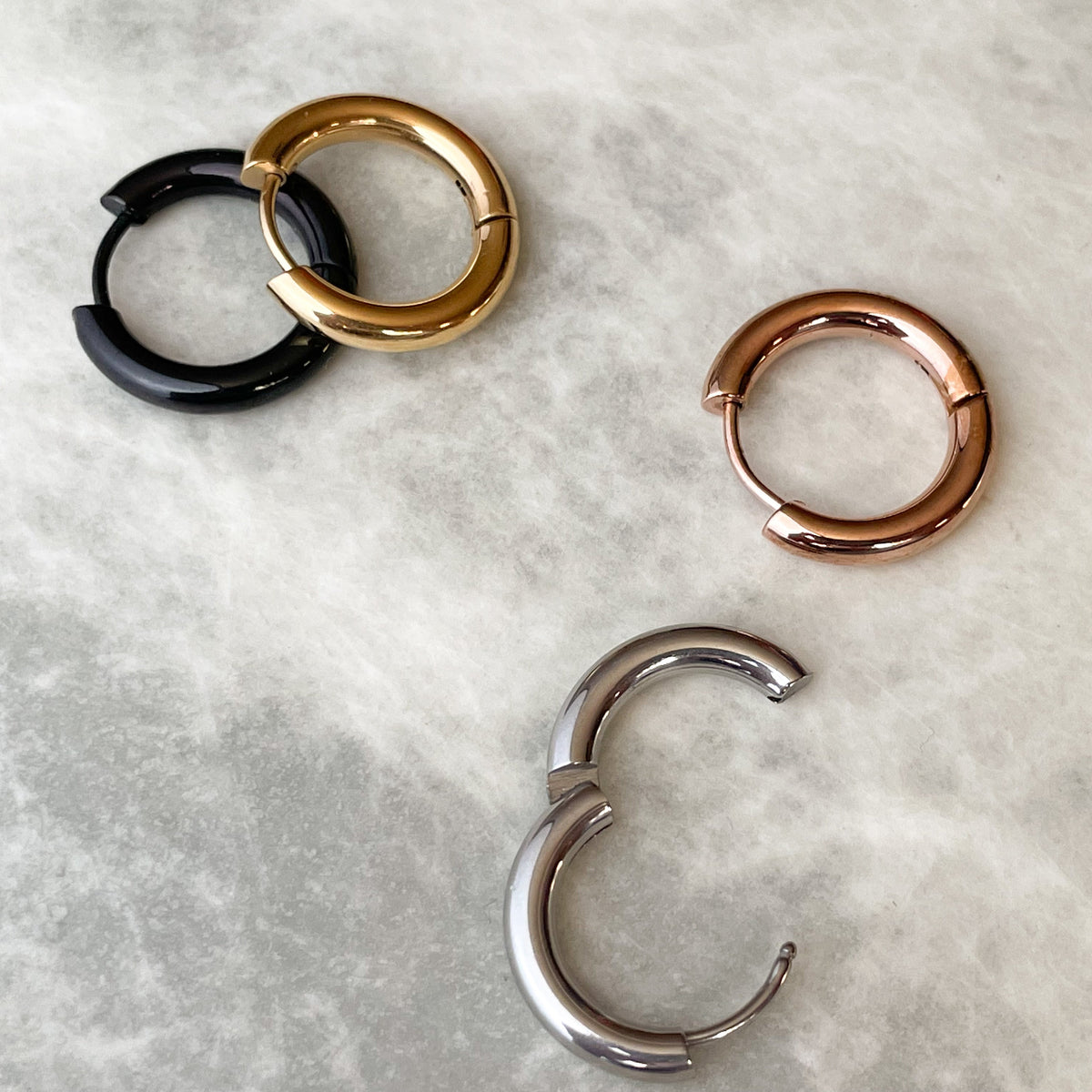 Four hoop earrings in black, gold, rose gold, and silver , with the silver one open showcasing the hinged closure, on a light gray marble surface.