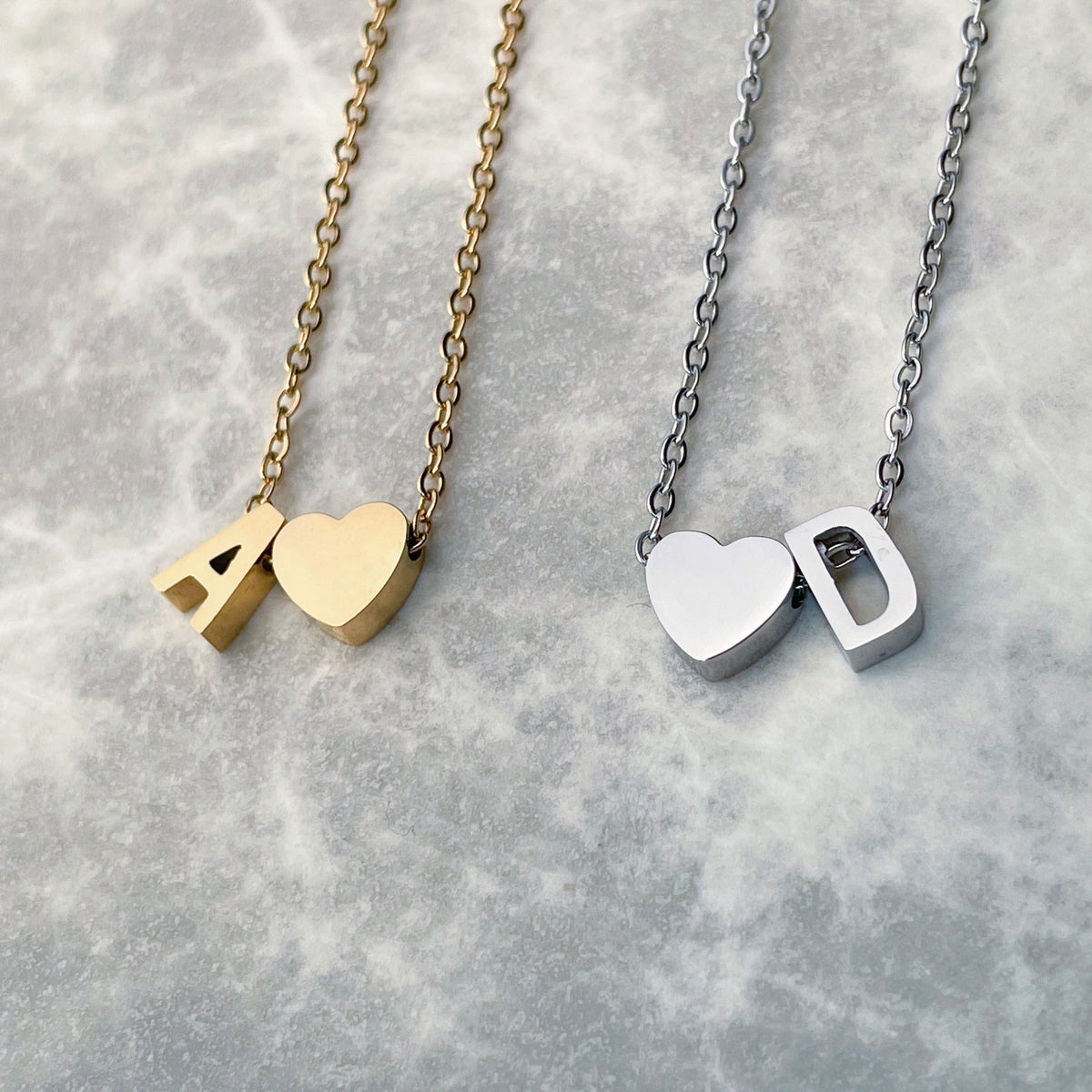 Two heart & initial charm necklaces, one gold and one silver, on a textured gray surface.