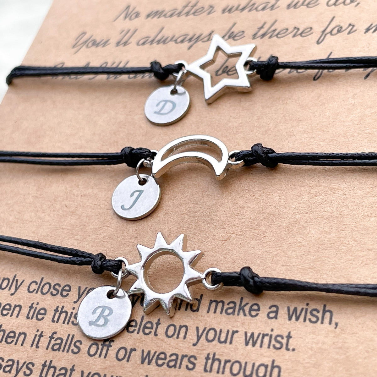 Three black cord bracelets with silver star, crescent moon and sun charms with initial discs on a textured surface.