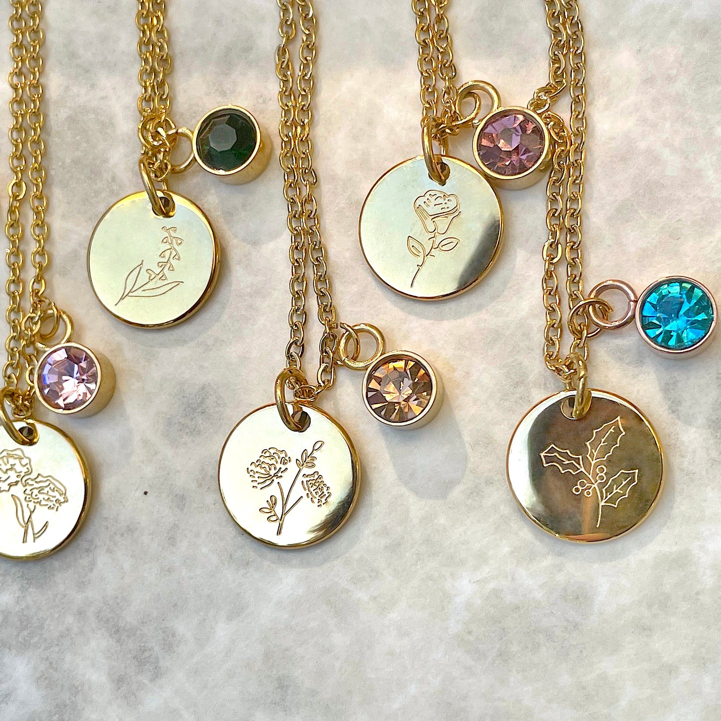 Close-up view of gold birth month necklaces with engraved pendants and colorful crystals on a marble surface