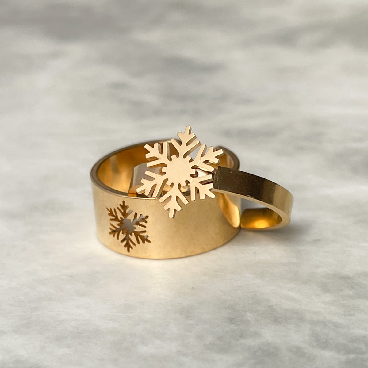 Two gold rings, one thin one wide with snowflake designs on a textured gray background