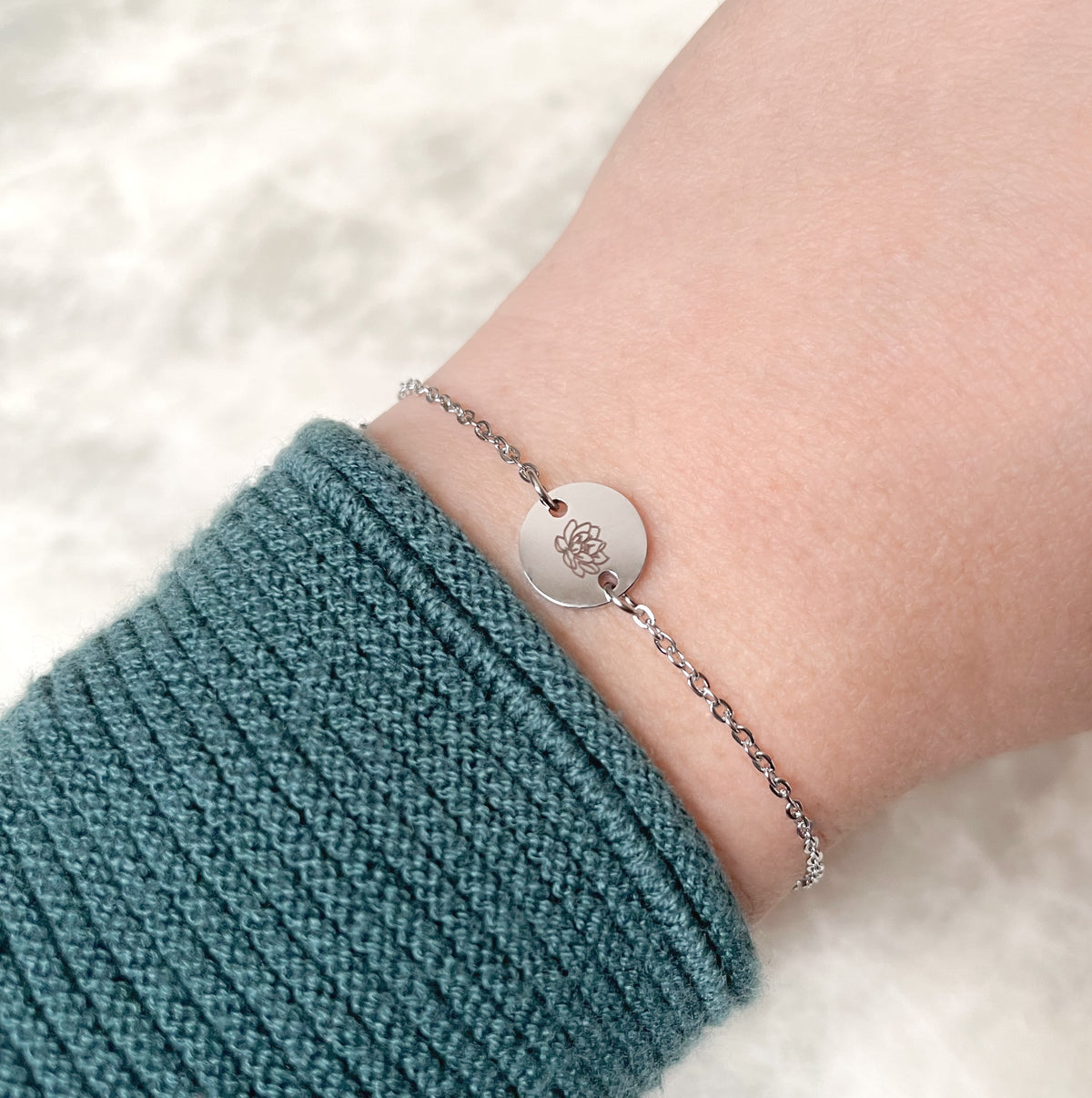 Silver bracelet with a lotus flower engraved round charm on a wrist wearing a teal sleeve against a white background