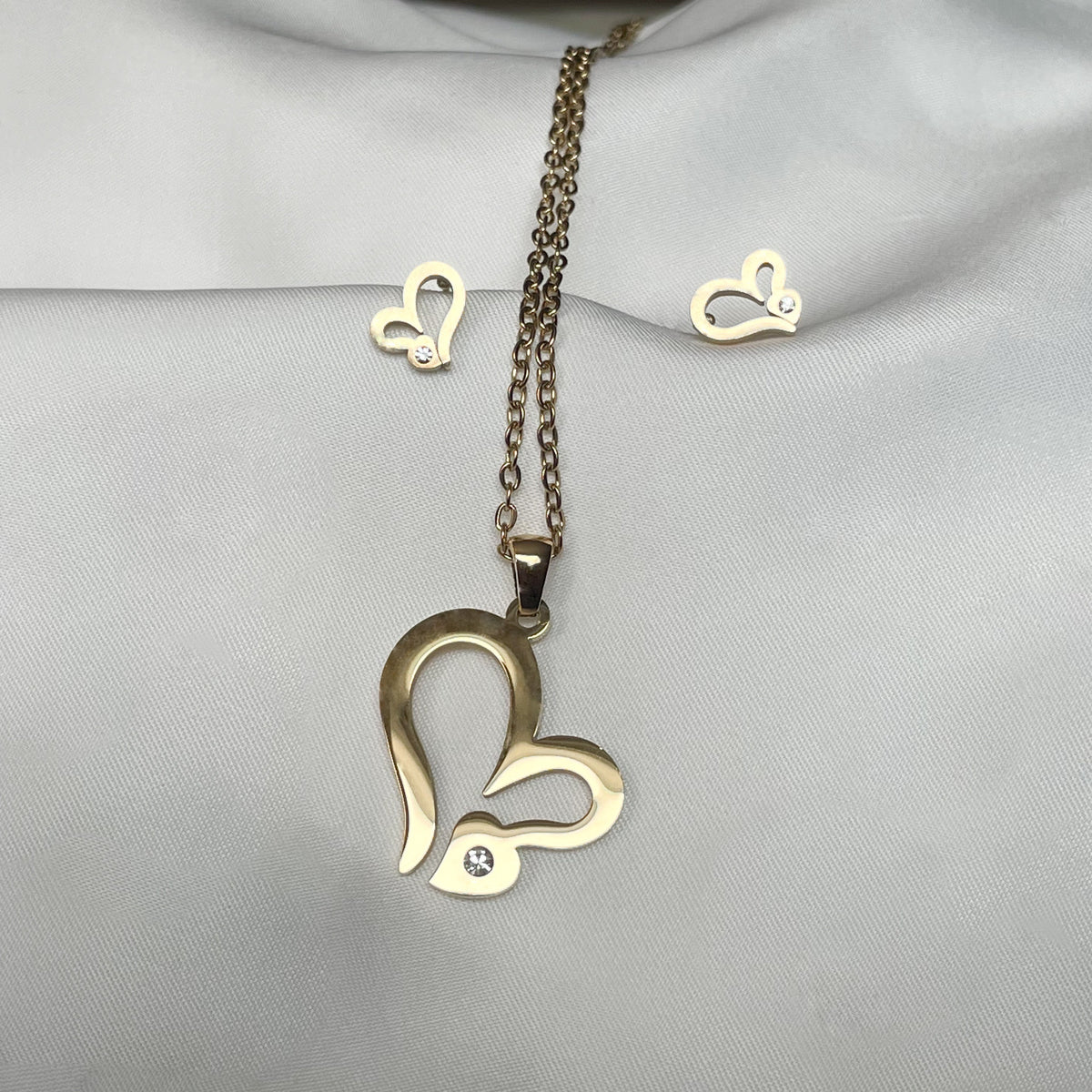 Large gold heart pendant necklace and matching stud earrings at the back each with a single CZ accent on a light gray fabric background