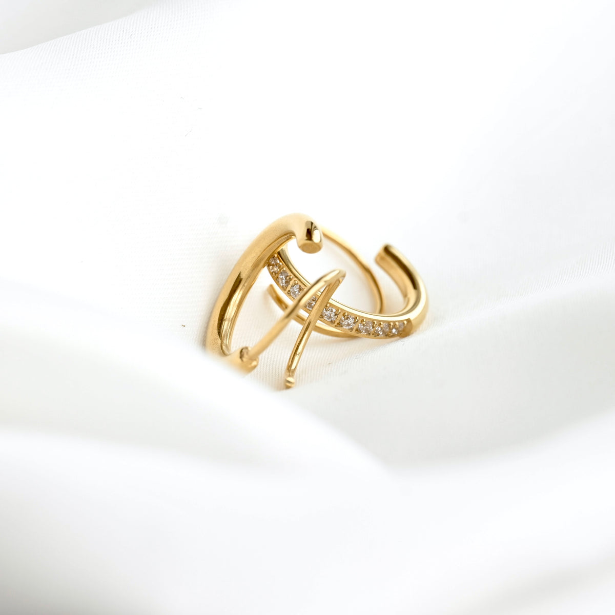 A gold huggie hoop ring with a curved bar studded with small clear crystals, resting on a soft white fabric background.