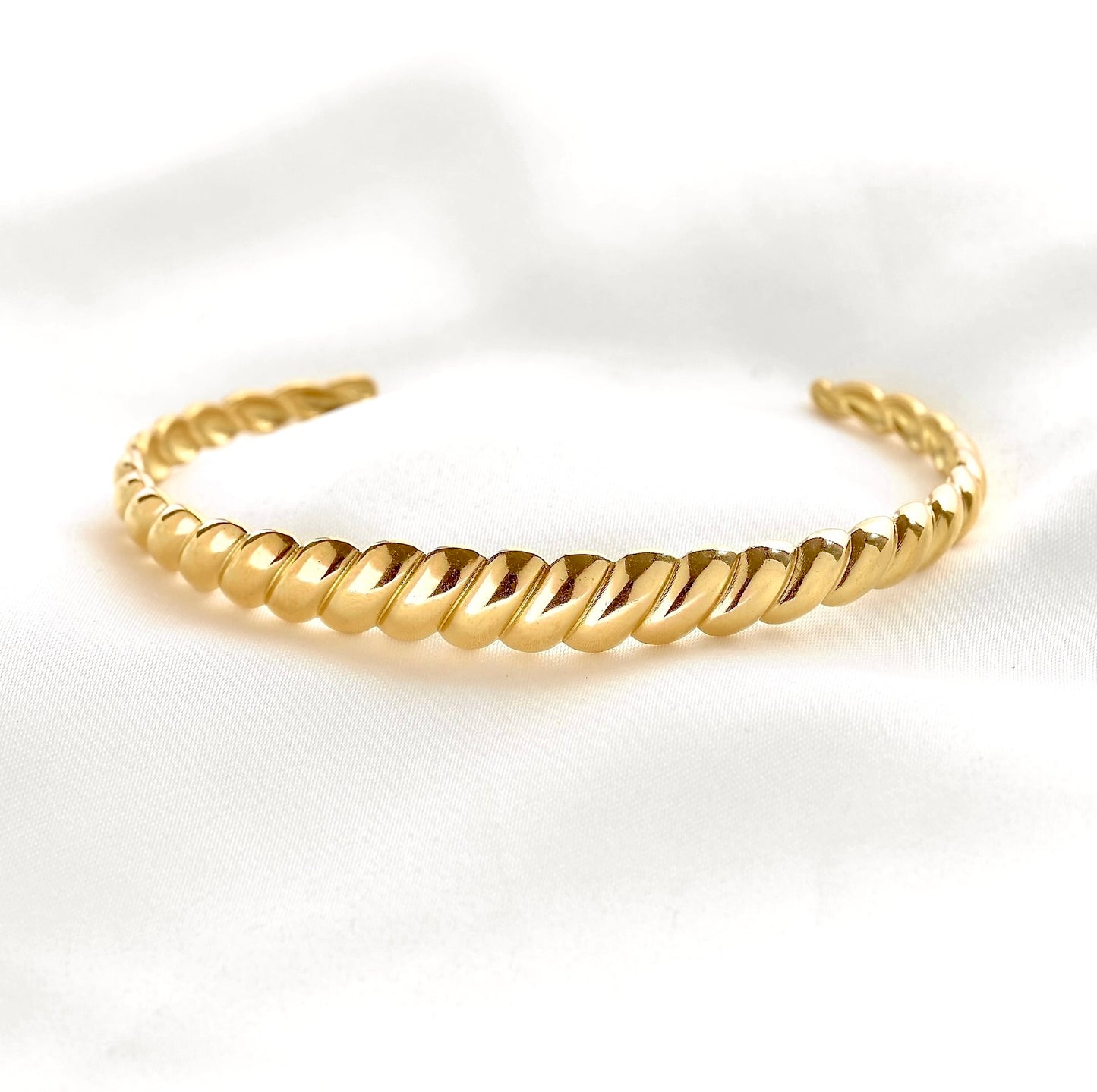 A gold-toned twisted rope cuff bracelet resting on a white textured fabric background.